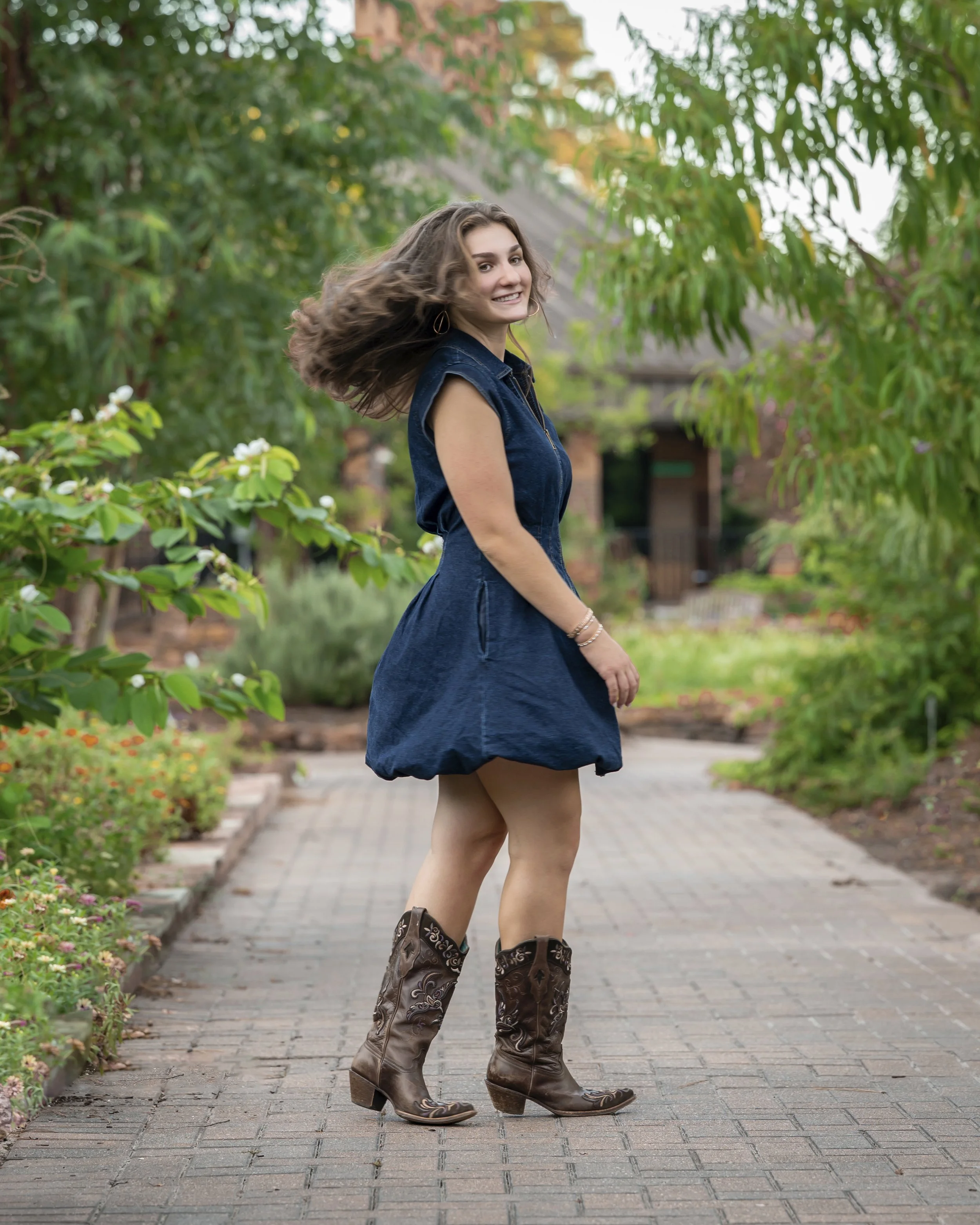 A young woman with curly brown hair smiling and looking over her shoulder while walking on a brick pathway in a garden, wearing a sleeveless denim dress and cowboy boots.