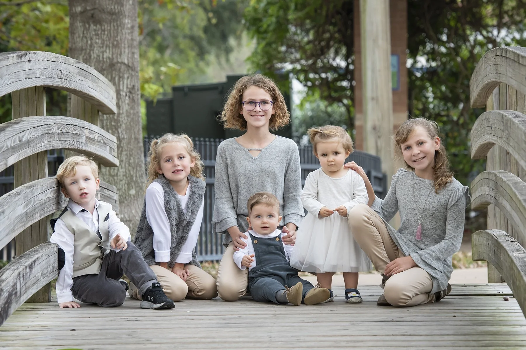 A group of seven children, three boys and four girls, outdoors on a wooden bridge, smiling and posing for a photo in a park with trees and greenery in the background.