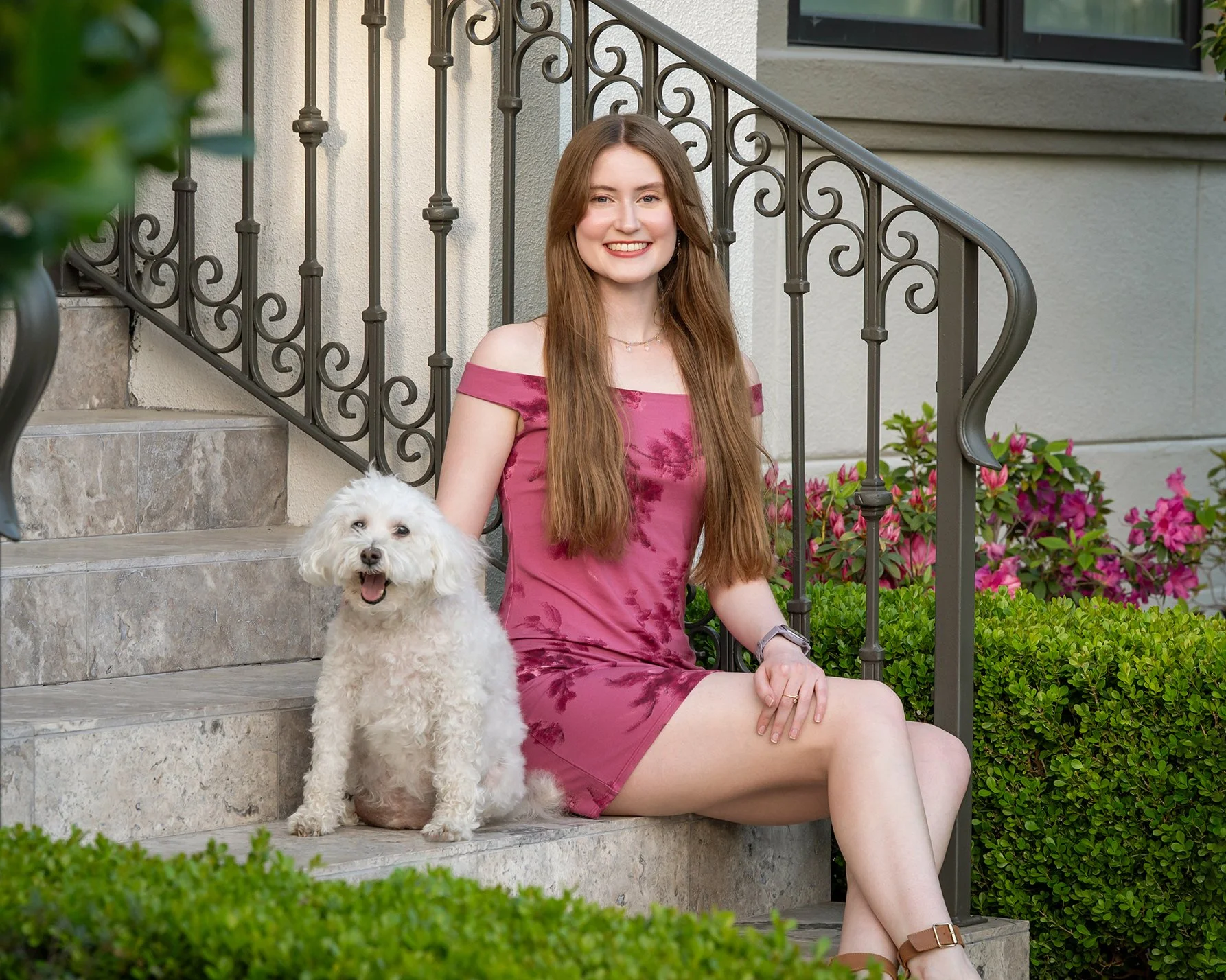 A young woman with long brown hair, wearing a pink off-shoulder dress, sitting on stairs outside of a house, smiling, with a small white dog sitting beside her.