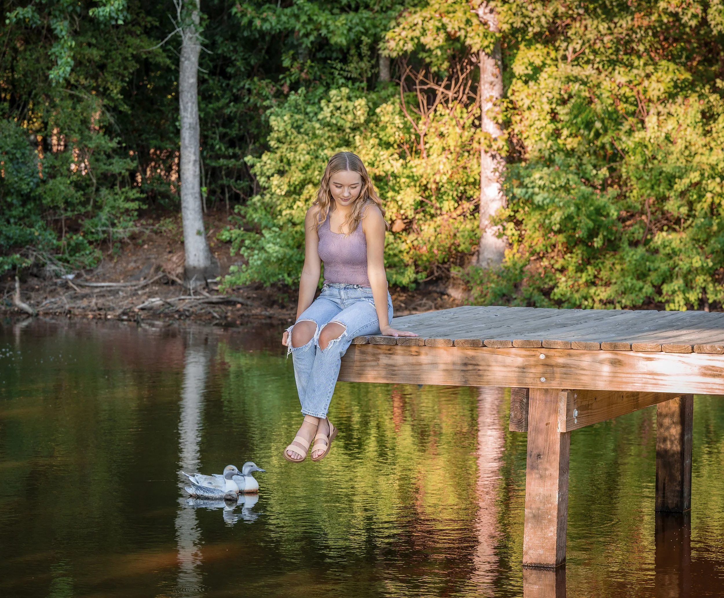 A senior girl with long wavy hair sitting on a wooden dock over a lake, with trees and colorful foliage in the background. Two ducks are swimming in the water near the dock.