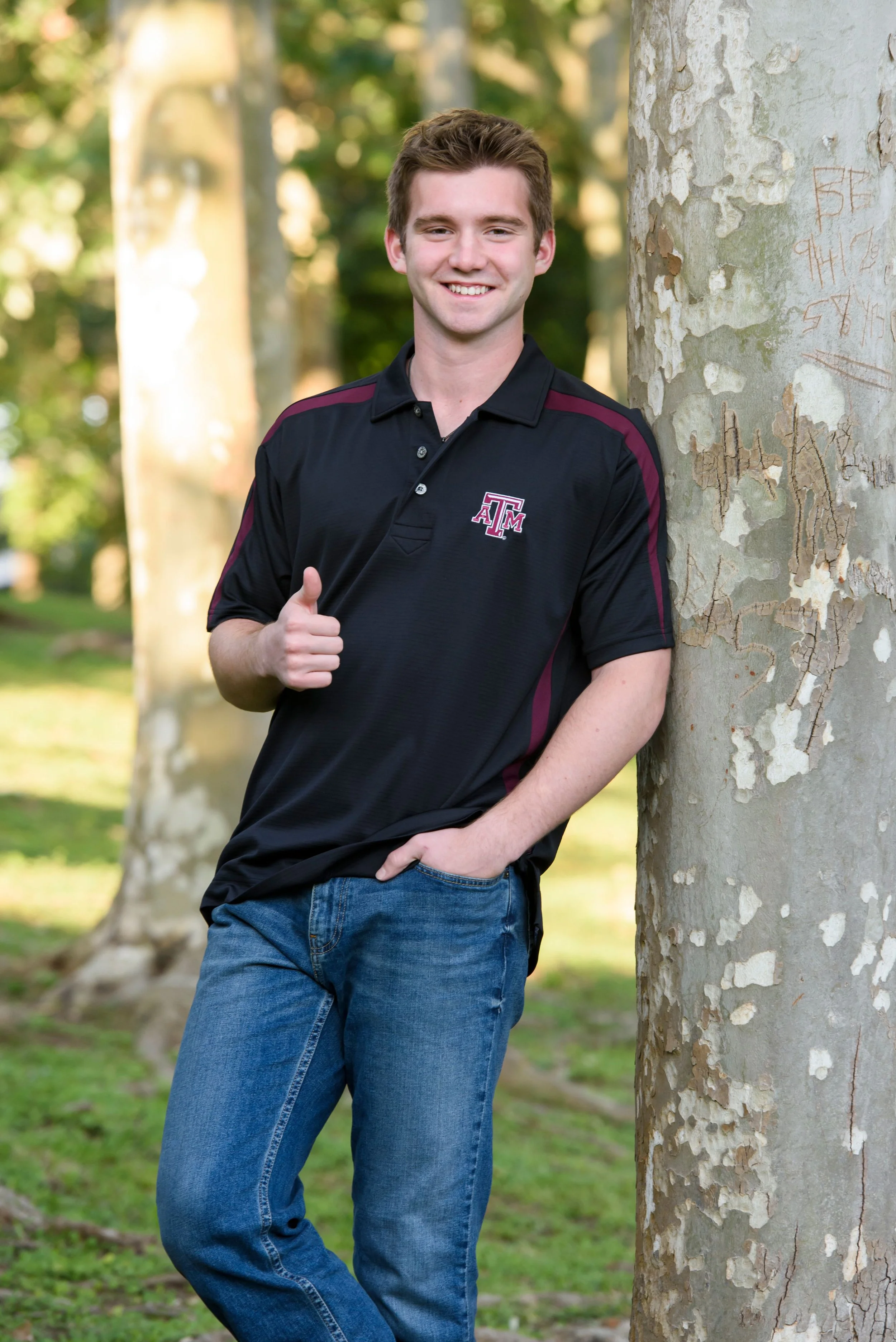 A senior guy with short brown hair, smiling and giving a thumbs-up, leaning against a tree in a park with green leaves in the background, wearing a black Texas A&M University polo shirt and blue jeans.
