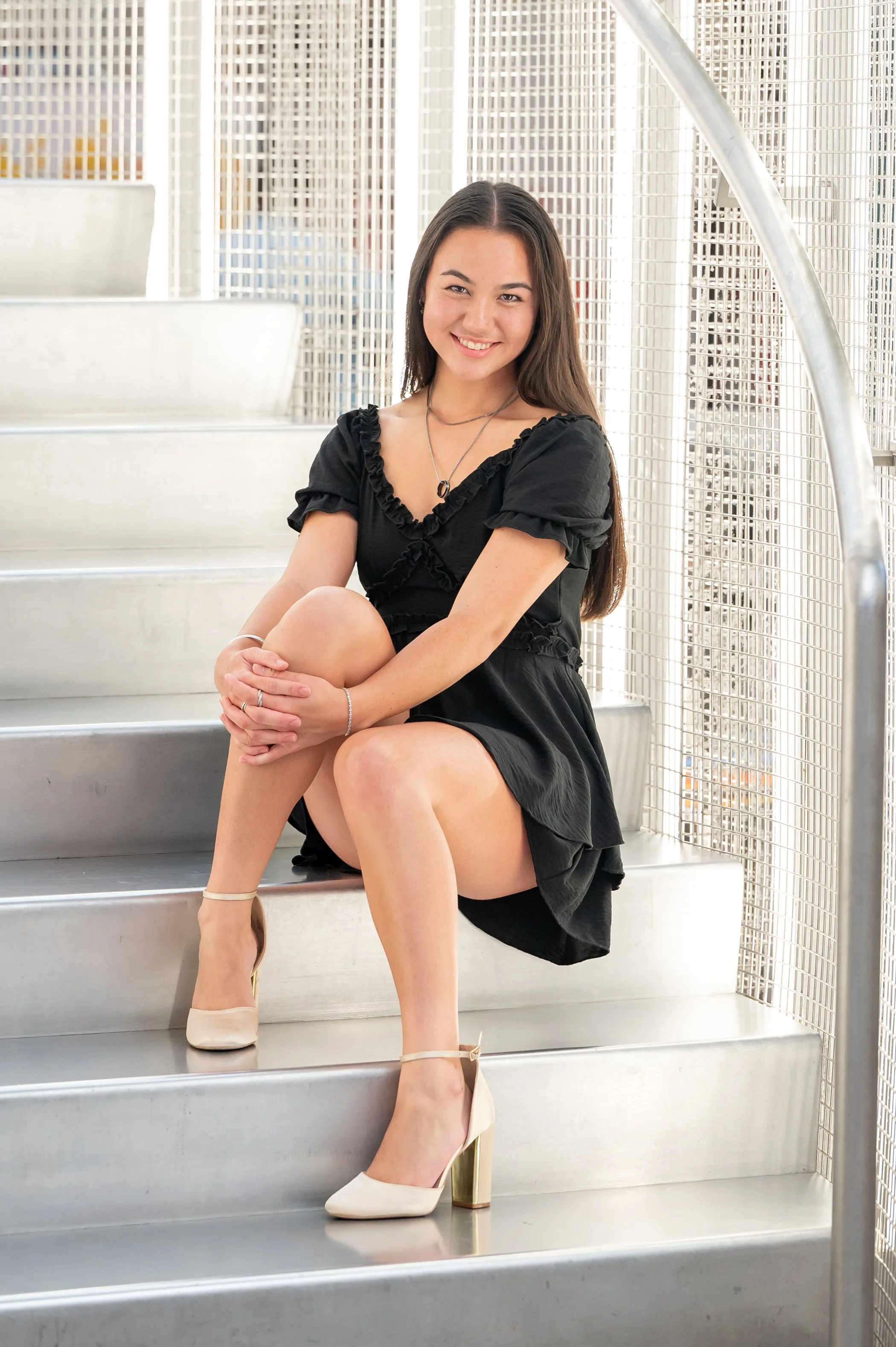 Portrait of a smiling senior girl sitting on metal stairs in front of a metallic mesh railing in a modern building.