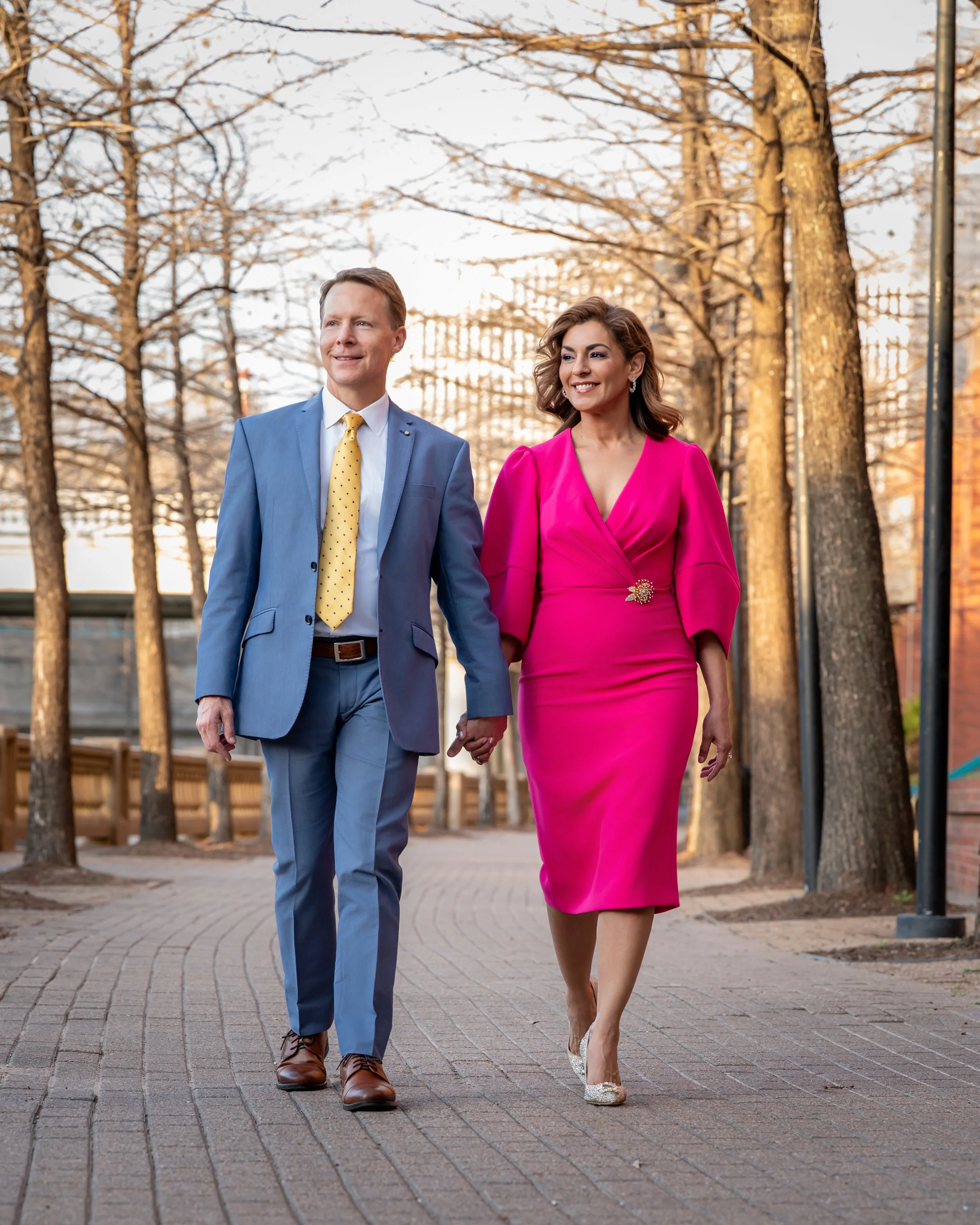 A well-dressed couple walking hand-in-hand outdoors in a park with leafless trees, during possibly late fall or winter, enjoying a sunny day.