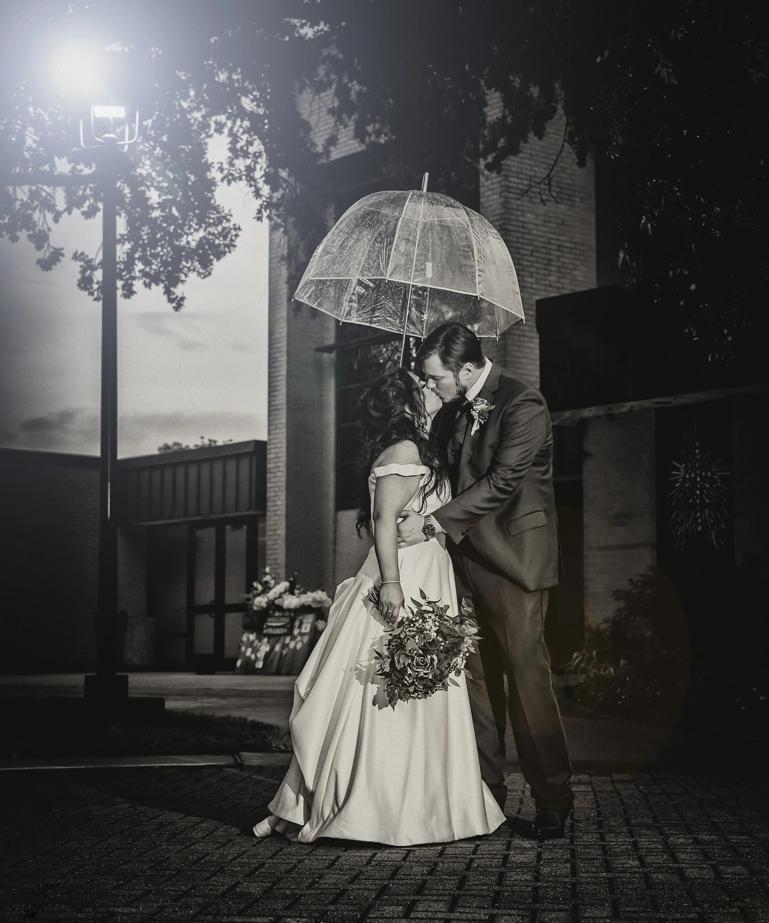 A couple sharing a kiss on a rainy night, with the woman holding a bouquet of flowers, under a transparent umbrella, illuminated by a streetlamp.