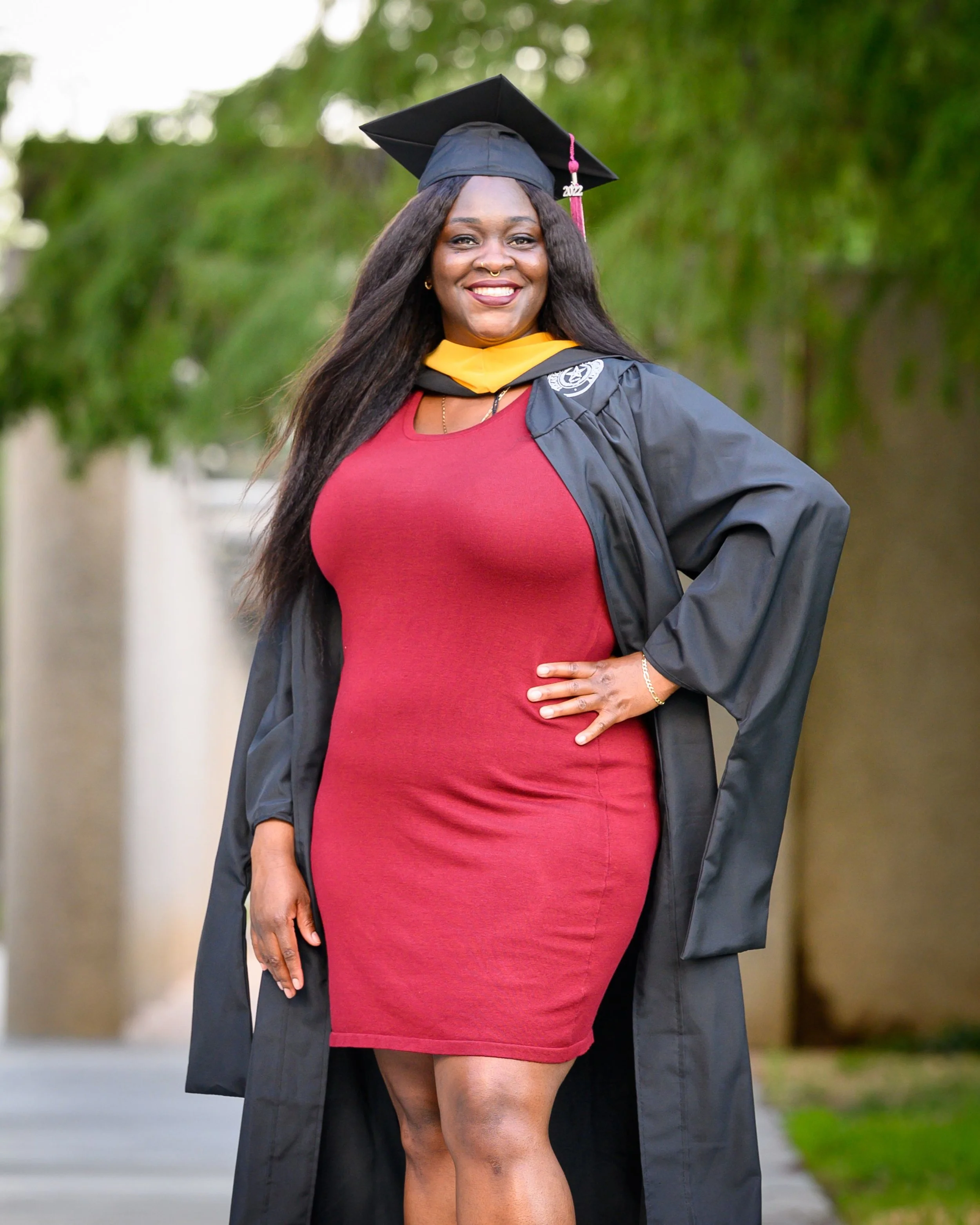 Woman celebrating graduation outdoors in a red dress and black graduation gown with a mortarboard cap, smiling and posing confidently.
