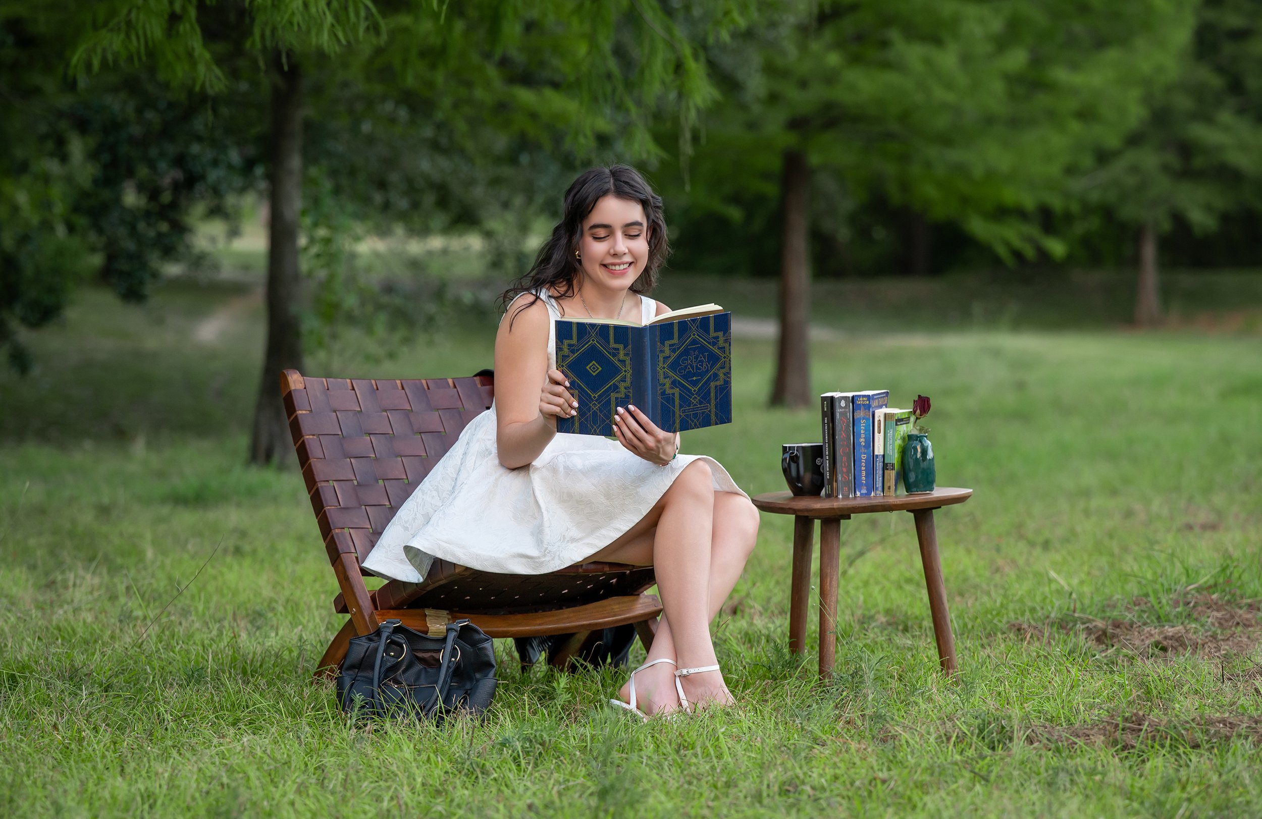 A smiling woman in a white dress sits on a wooden outdoor lounge chair, reading a book titled 'The Great Gatsby' in a lush green park with trees in the background. A small side table holds additional books, a camera, and a green vase. A black bag is 