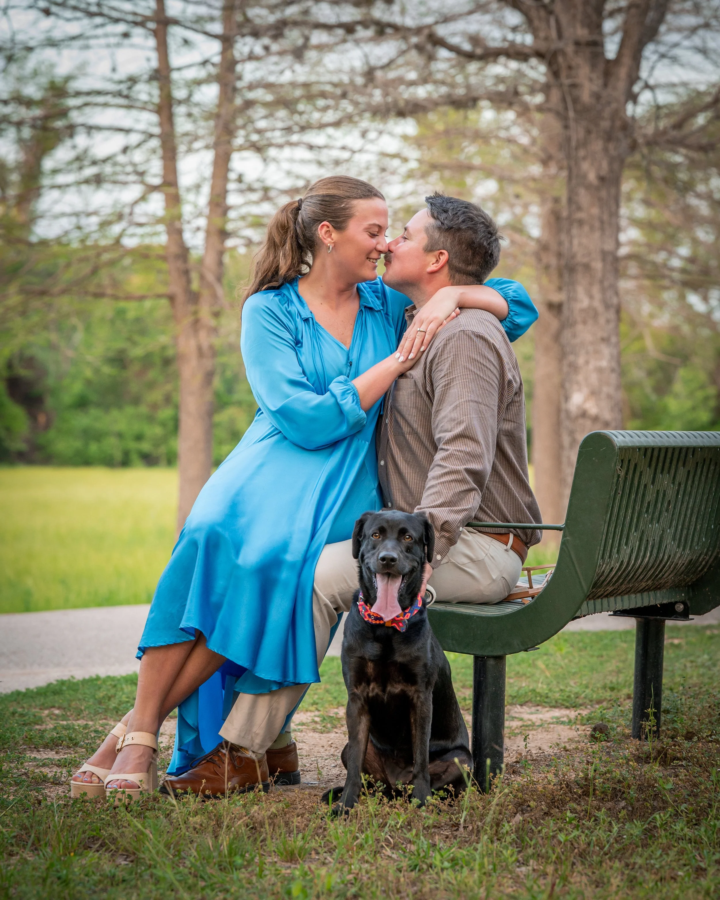 A woman and a man are close together, nose to nose, smiling in a park with trees in the background. The woman is wearing a blue dress and the man is seated on a park bench. There is a black dog sitting in front of the man, looking at the camera, with