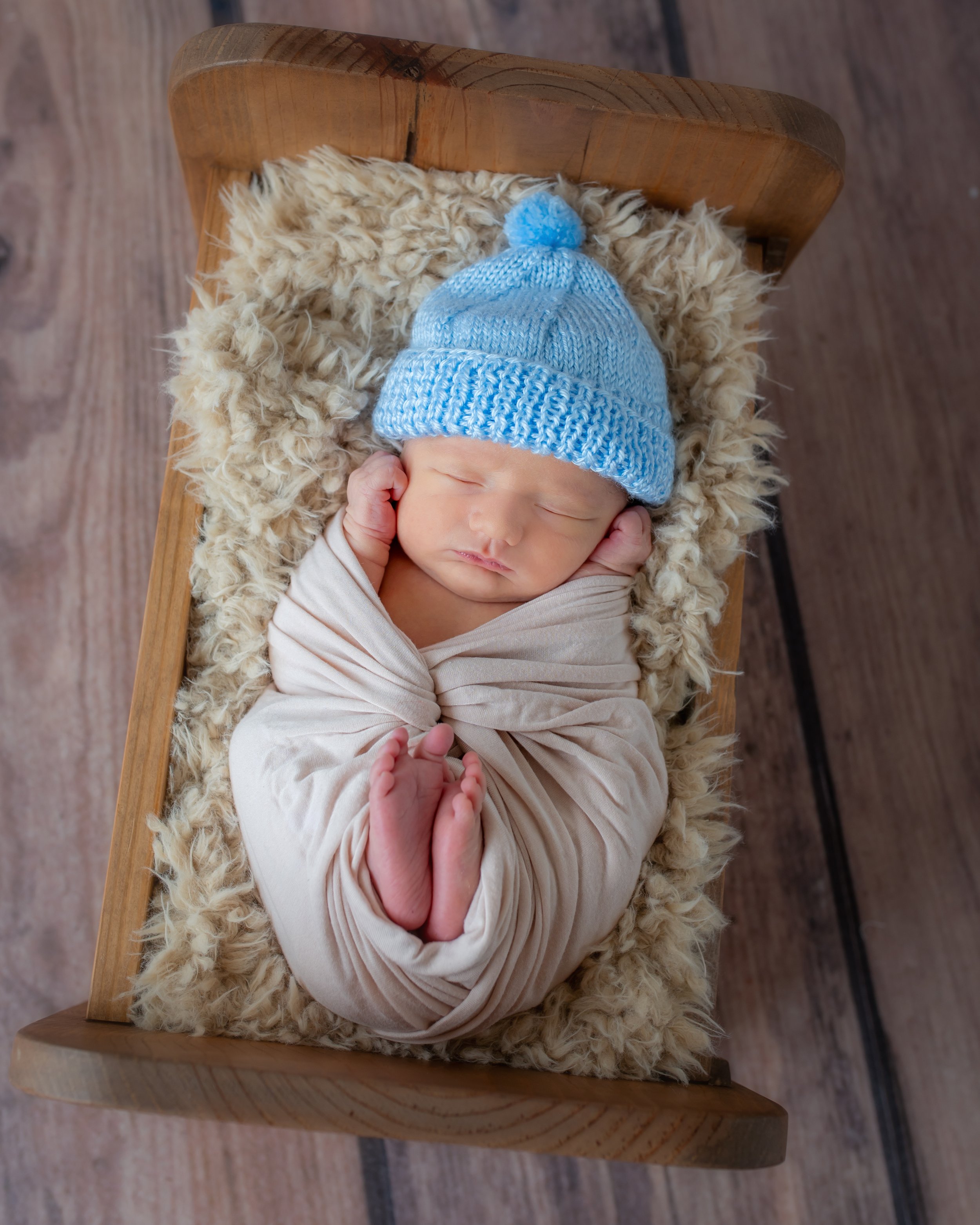 A newborn baby lying in a wooden cradle, wearing a gray knit hat with a pom-pom. The baby is wrapped in a beige blanket with a fluffy cream-colored lining, peaceful and resting.