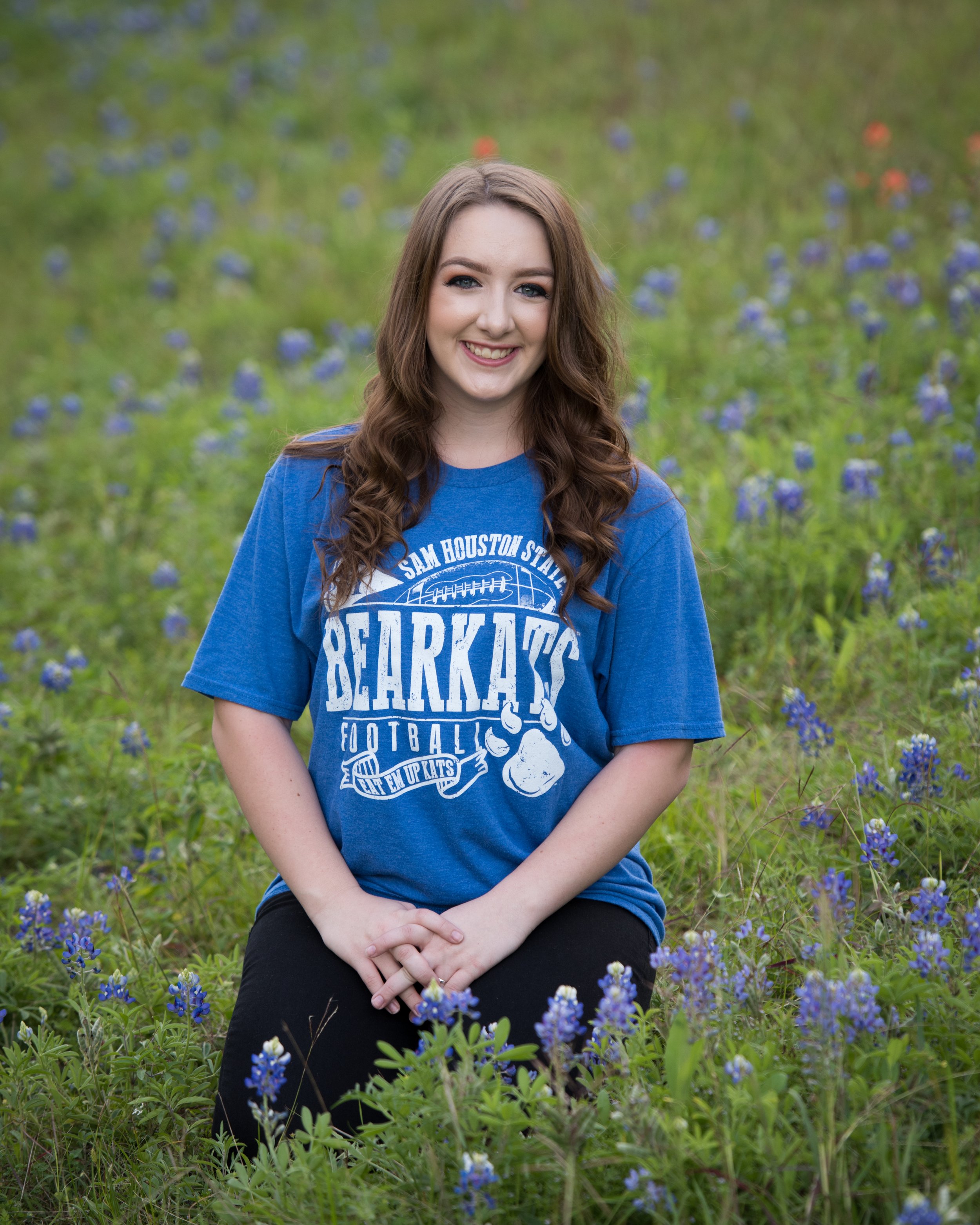 A senior girl with long, wavy brown hair, wearing a blue T-shirt with a college team logo, sitting outdoors in a field of bluebonnet flowers, smiling at the camera.