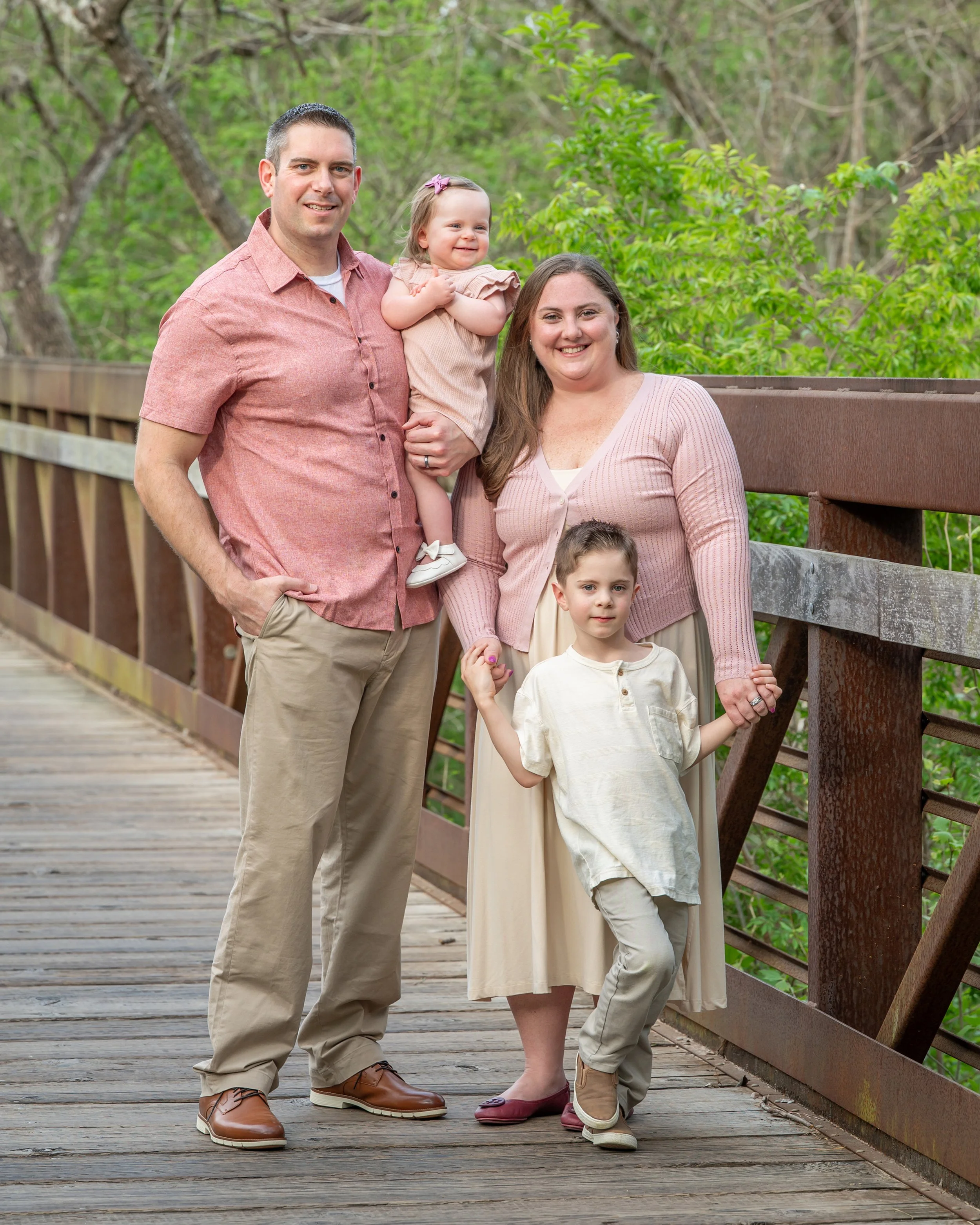 A family of four, including a man, a woman, a young girl, and a boy, posing on a wooden bridge outdoors surrounded by greenery.