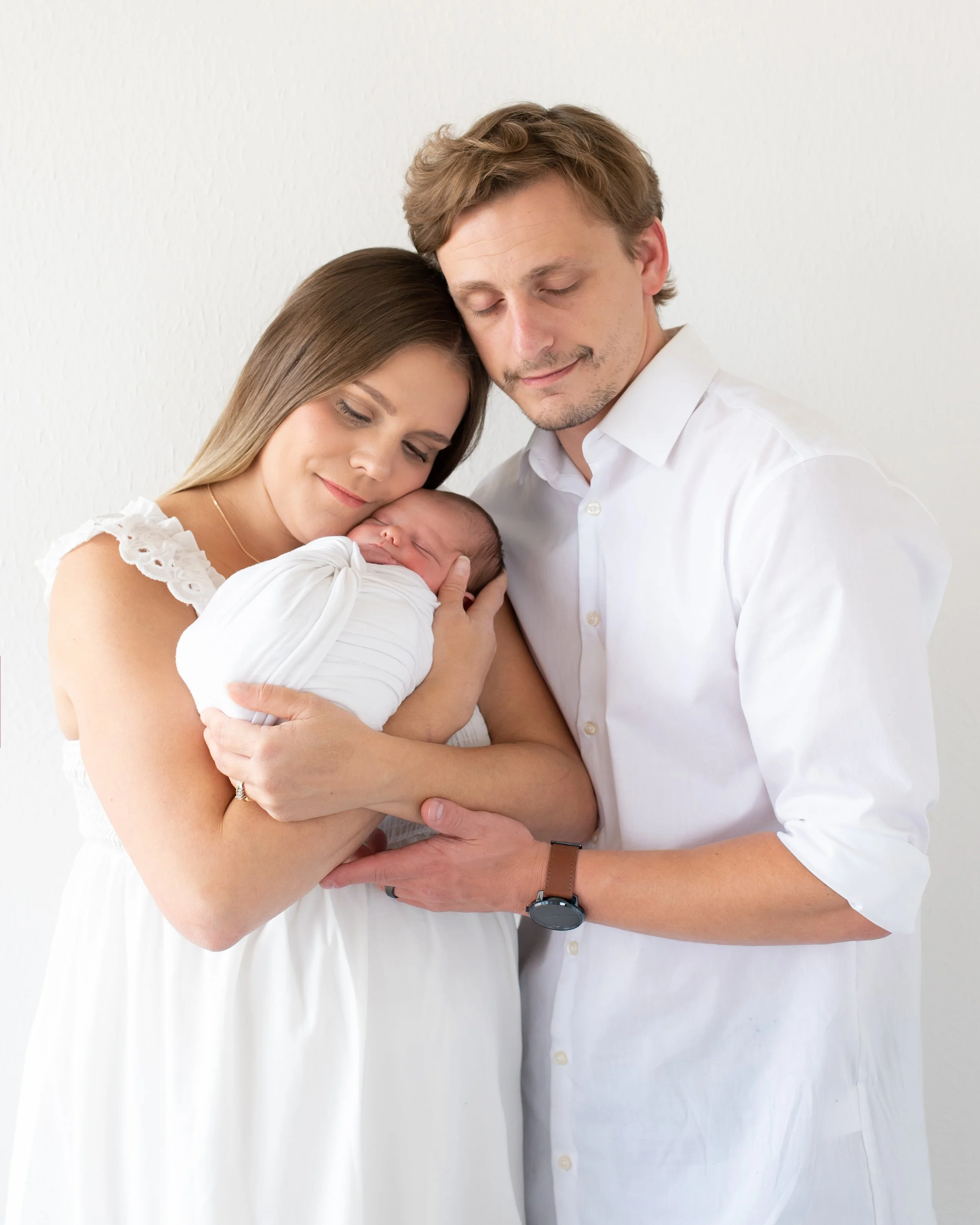 A proud family photo of a young couple with their newborn baby. The woman has long brown hair, and the man has light brown hair. They are both wearing white shirts and are hugging their sleeping infant.
