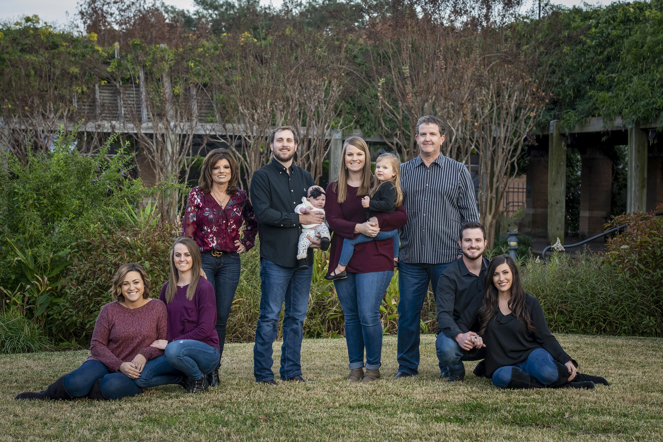 A large group of people, including men, women, and children, posing outdoors in front of trees and bushes during daytime.