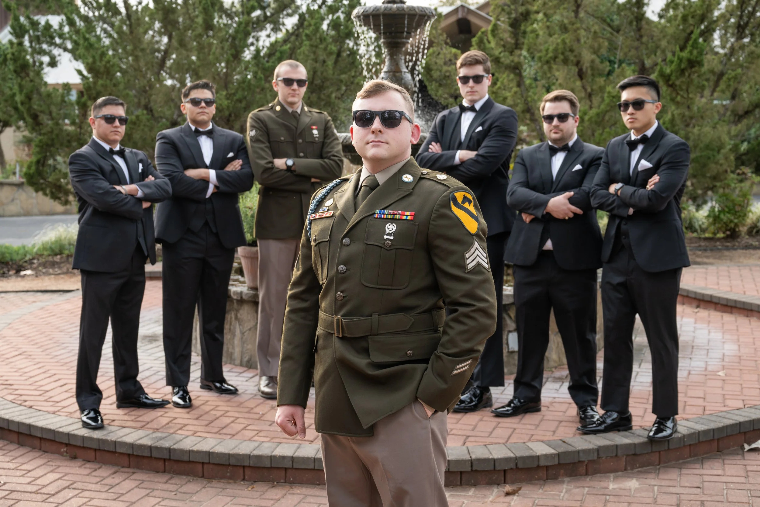 A group of seven men, six dressed in tuxedos and sunglasses, standing with arms crossed around a fountain featuring water cascading into a basin, in an outdoor setting with trees and brick paving.
