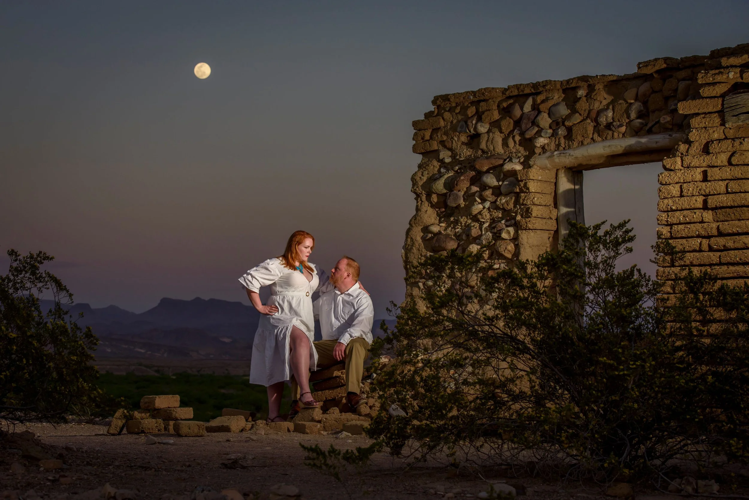 A woman and a man in white clothing are outdoors at dusk, with the full moon visible in the sky. The woman is standing with her hands on her hips, and the man is kneeling on one knee, looking at her. Behind them, there is a stone and brick structure 