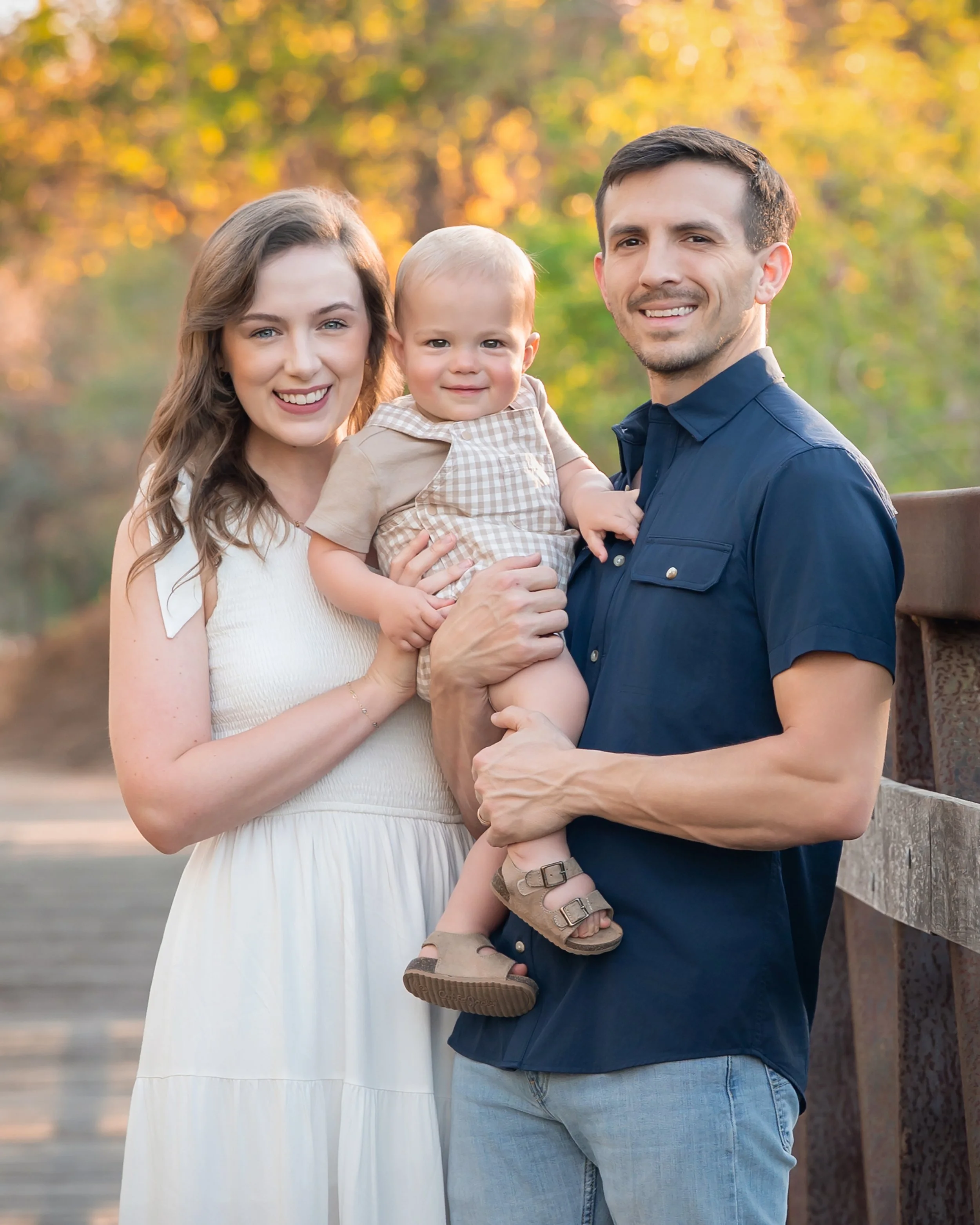 A happy family of three, a woman, a man, and a young child, standing outdoors at sunset with fall foliage in the background. The woman is wearing a sleeveless white dress, the man is dressed in a navy blue shirt and jeans, and the child is dressed in