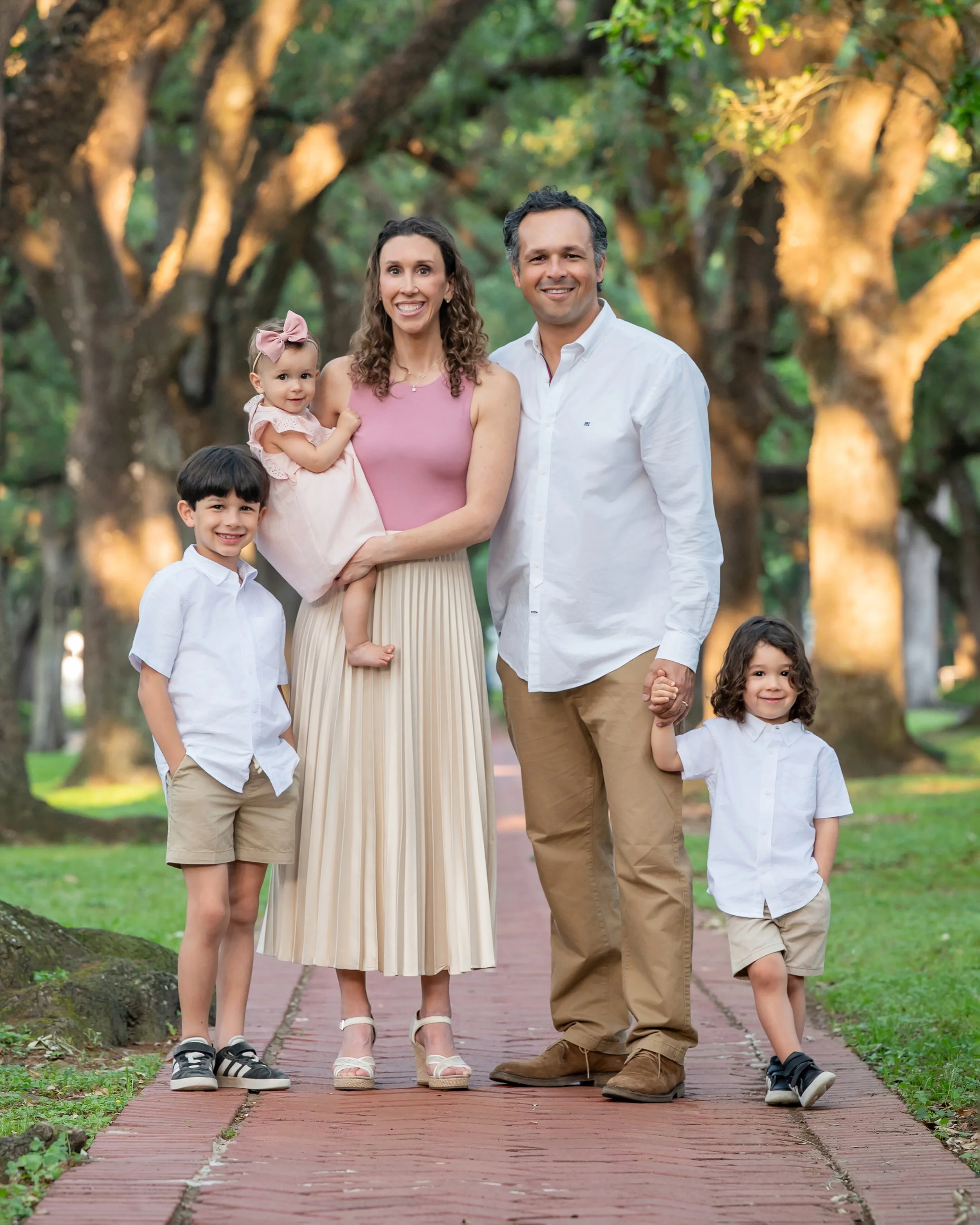 A smiling family of five, including mother, father, a girl, a boy, and a toddler girl, walking on a brick pathway in a park with green trees and sunlight in the background.