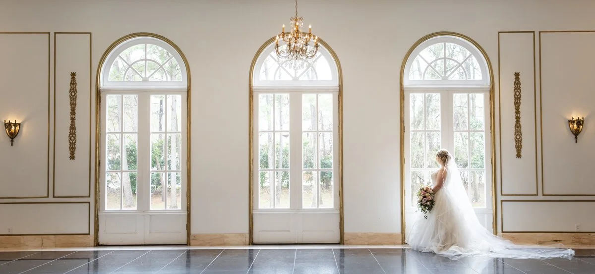 A bride in a white wedding dress holding a bouquet, standing by a large window in an elegant room with gold trim and a chandelier.