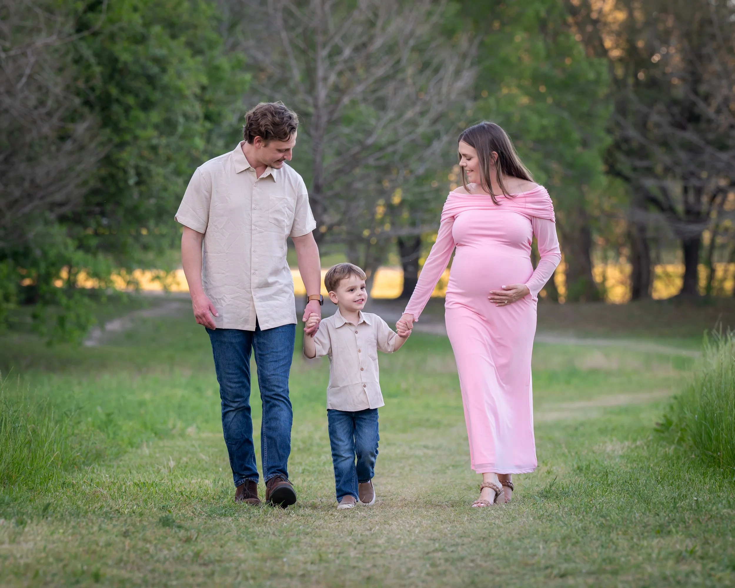 A pregnant woman in a pink dress walking hand-in-hand with a young boy while a man in beige shirt walks beside them in a park with green trees.