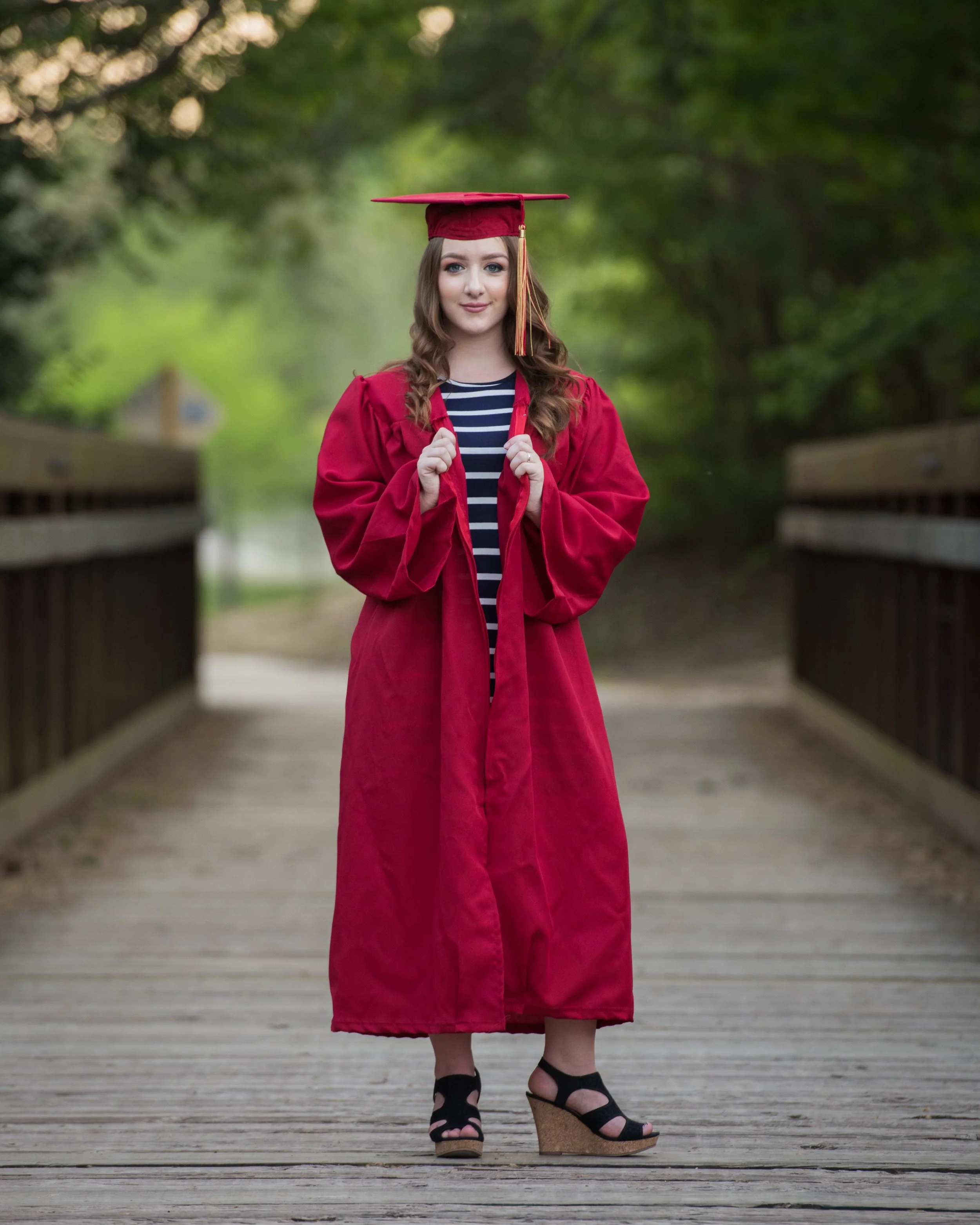 A young woman in a red graduation gown and mortarboard cap with a gold tassel stands on a wooden bridge in a green, wooded outdoor setting. She is holding the edges of her gown with both hands and is wearing black platform sandals.