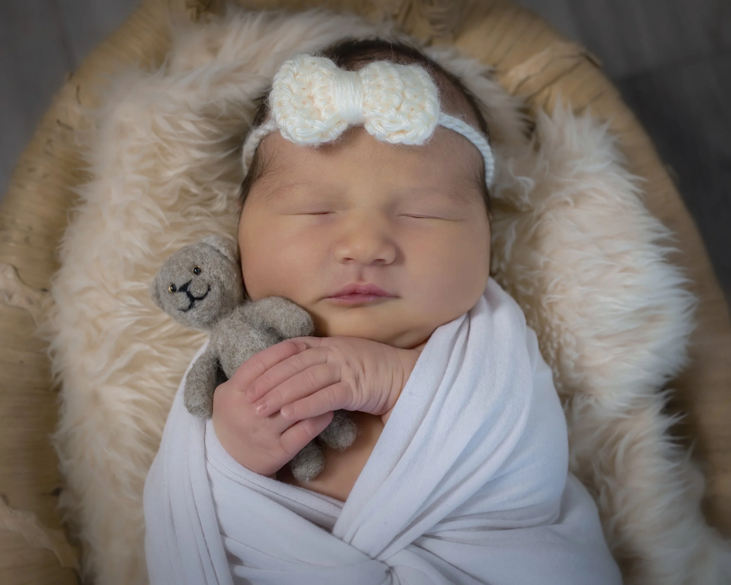 A sleeping newborn with a headband, cuddling a gray stuffed animal, lying on a fluffy cream-colored blanket.