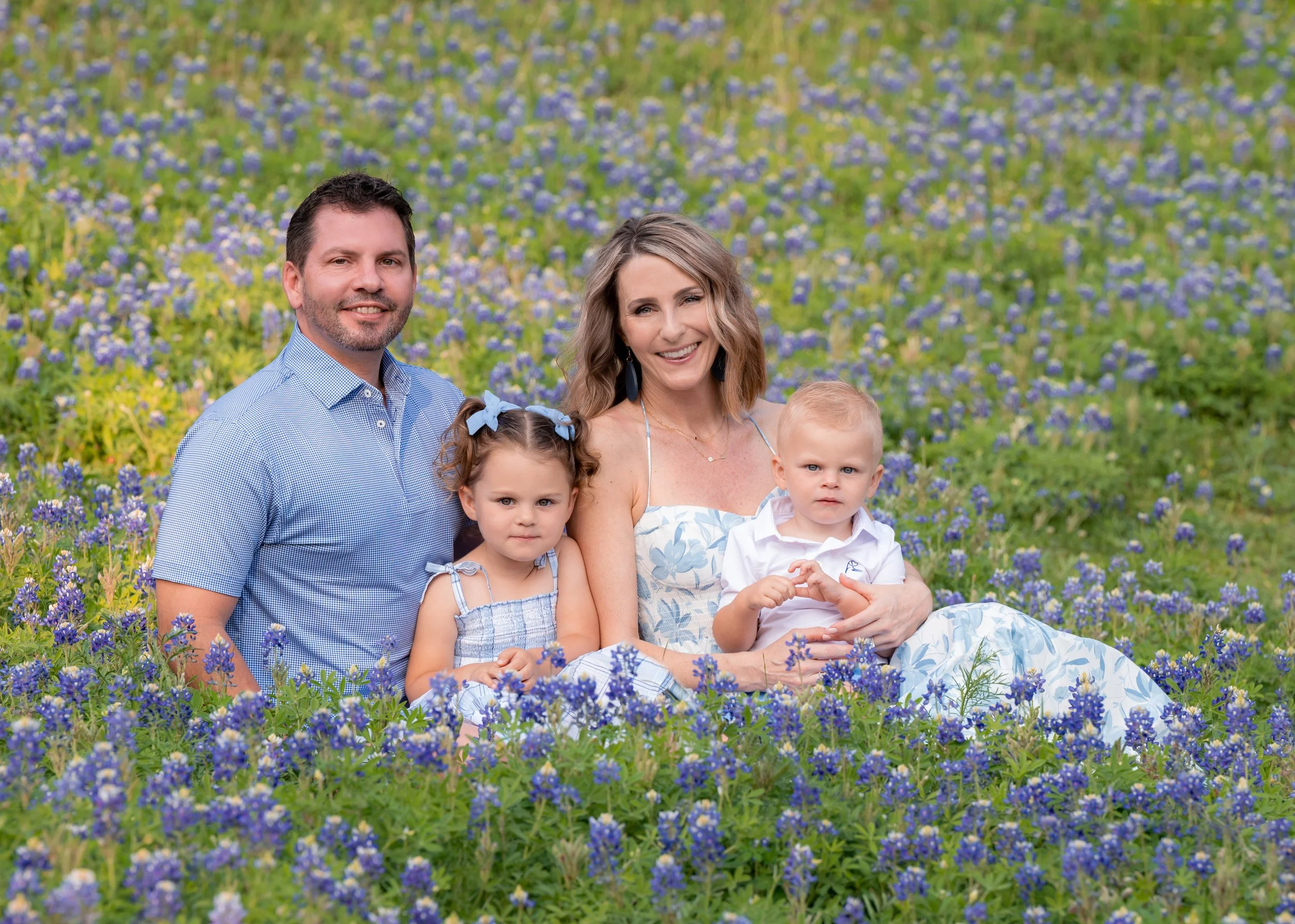 A family of five sitting on a field of bluebonnet flowers, smiling at the camera. The family includes two adults and three children, with a background of purple flowers and greenery.