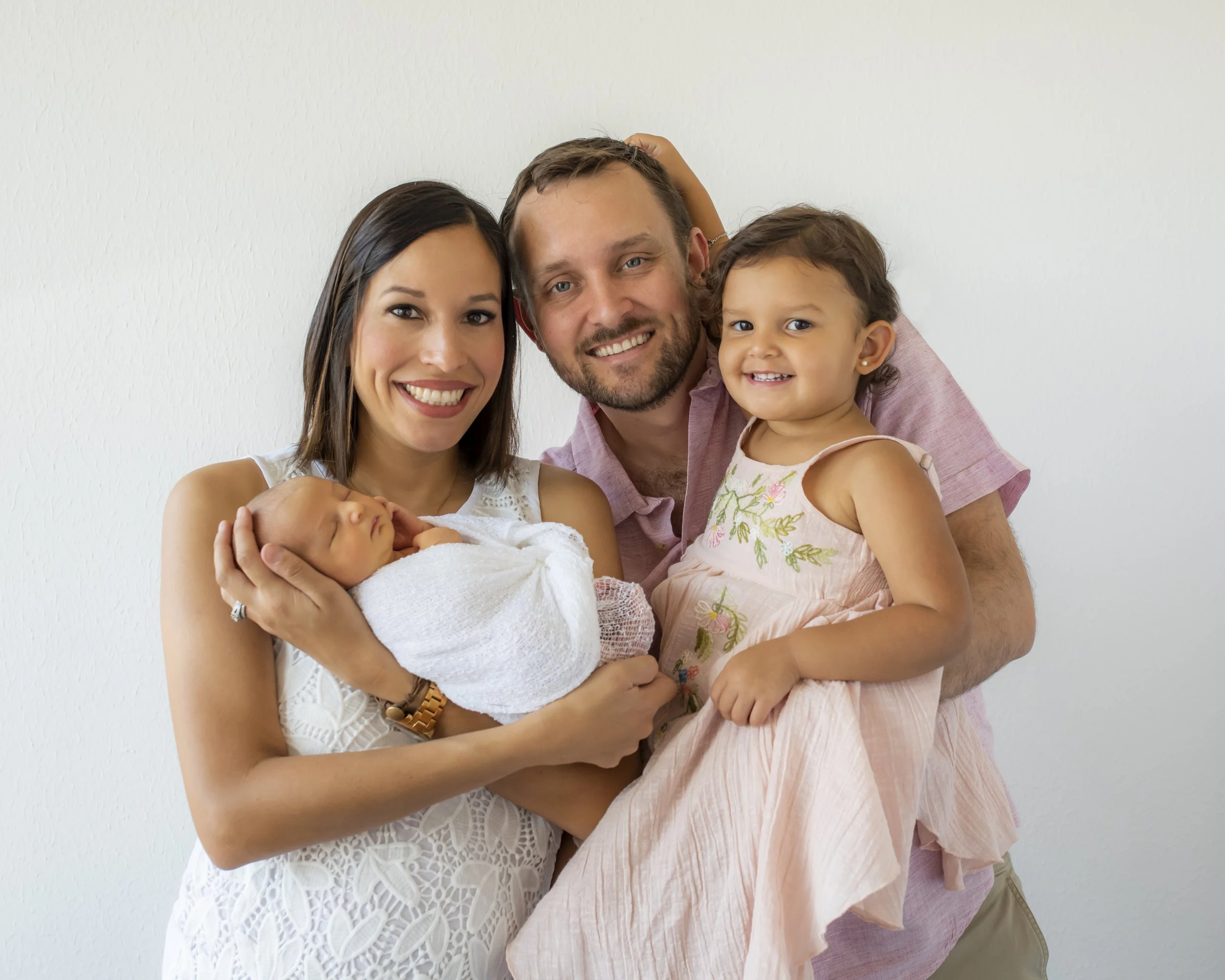 A happy family of four: a woman, a man, a young girl, and a newborn baby, posing together against a plain white wall. The woman is smiling and holding the baby wrapped in a blanket, the man is smiling with his arm around the girl, who is also smiling
