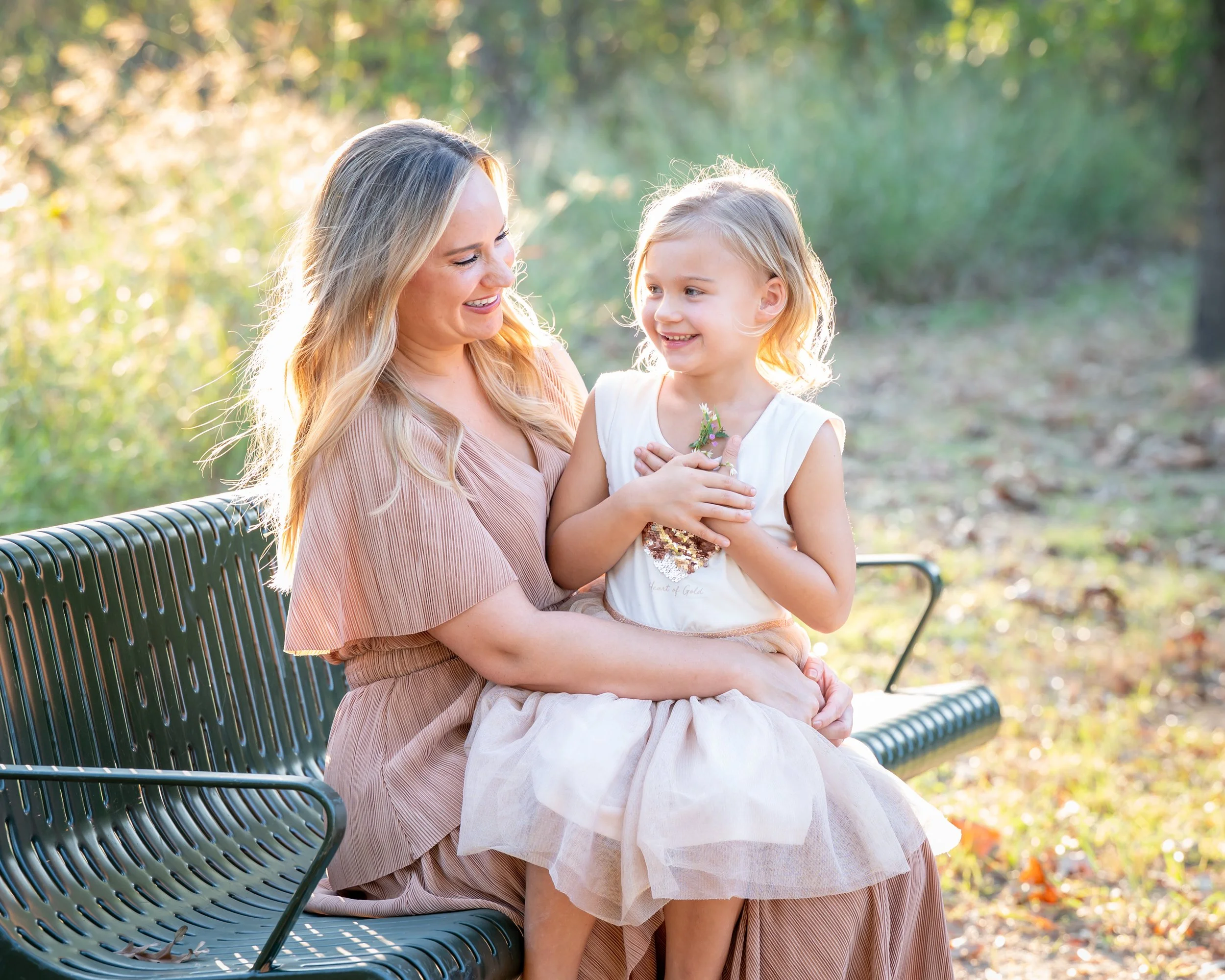A woman with long blonde hair and a young girl with blonde hair sitting on a park bench, smiling and looking at each other. The woman is wearing a pink dress, and the girl is wearing a white dress with a floral pattern. The background is outdoors wit