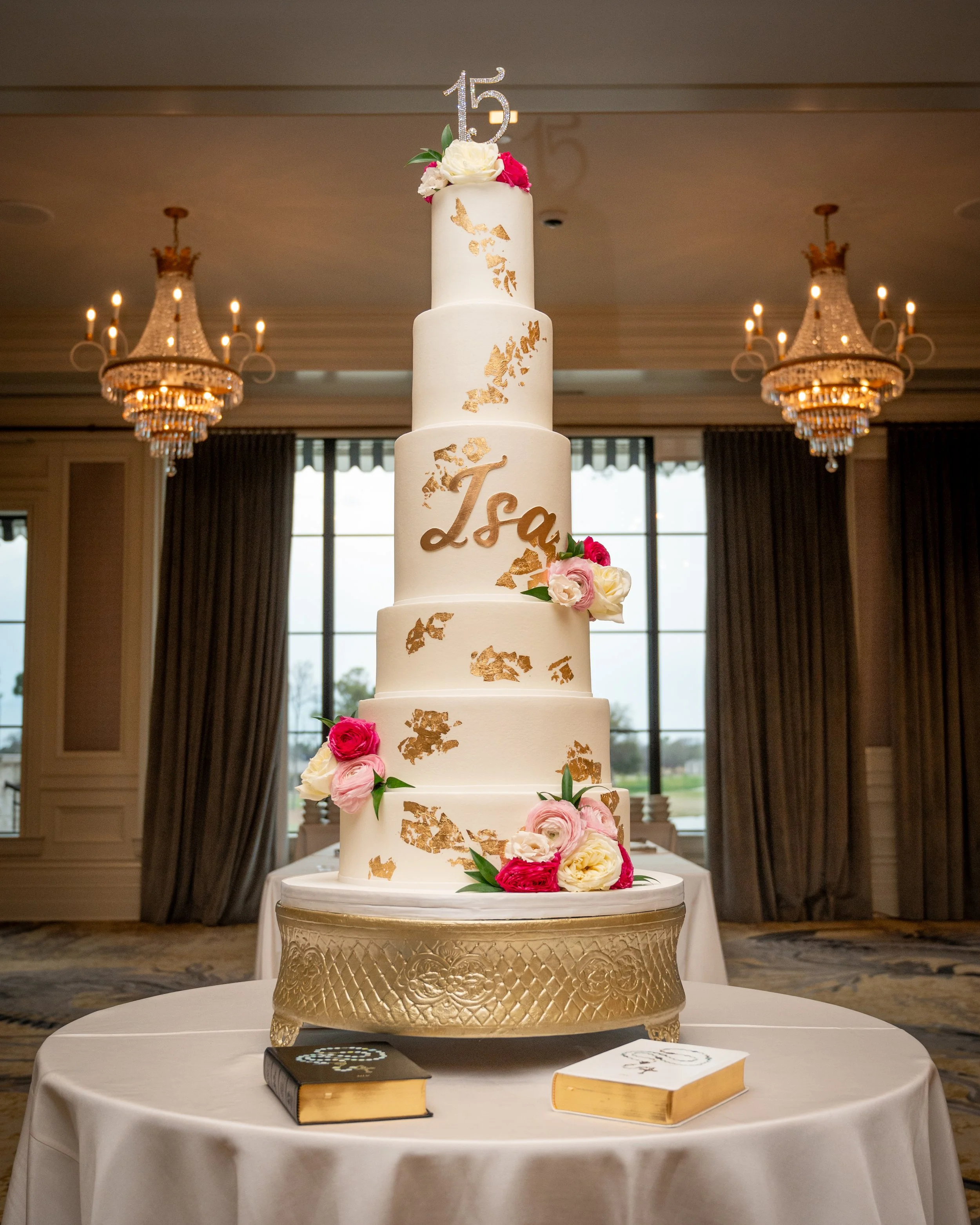 A tall, multi-tiered white wedding cake decorated with pink and white roses, gold leaf details, and a silver "15" topper for a quinceañera celebration, placed on a white table with two books underneath.