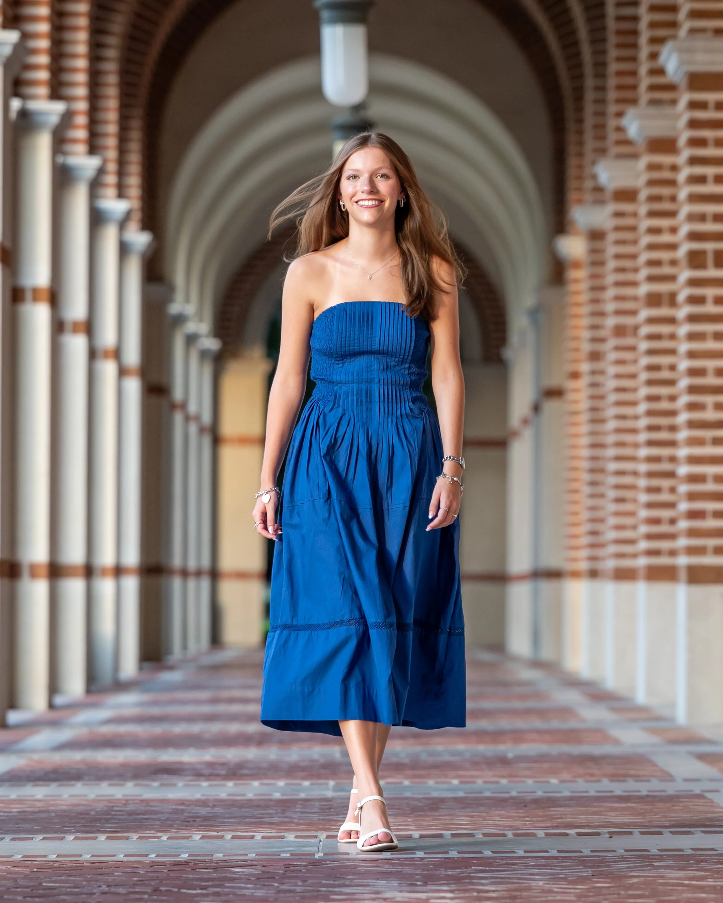 A woman with long brown hair wearing a blue strapless dress and white sandals walking in an arched corridor at Rice University with brick walls and columns, smiling.