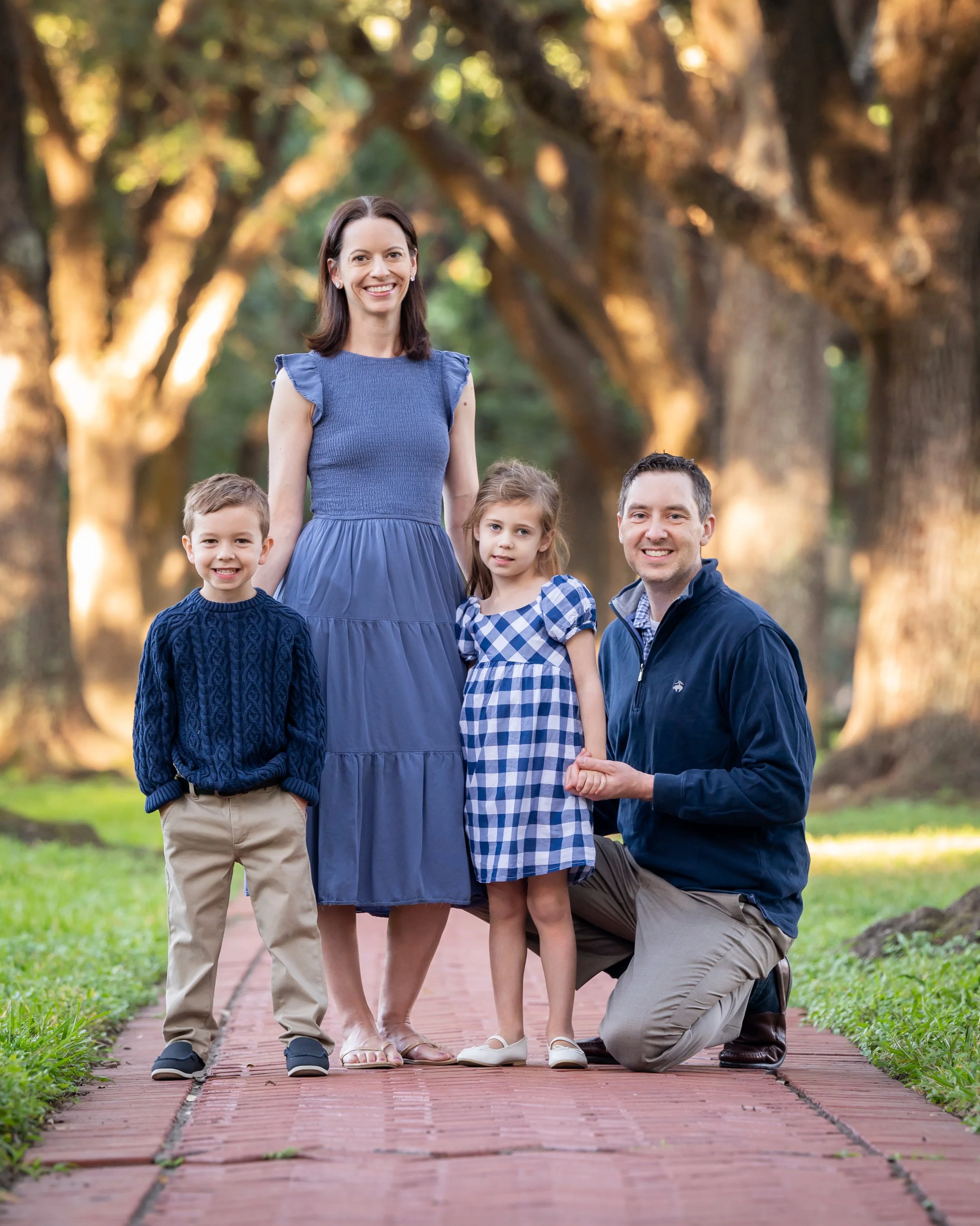 A family of four with a woman, man, son, and daughter posing outdoors on a brick path with trees and sunlight in the background.