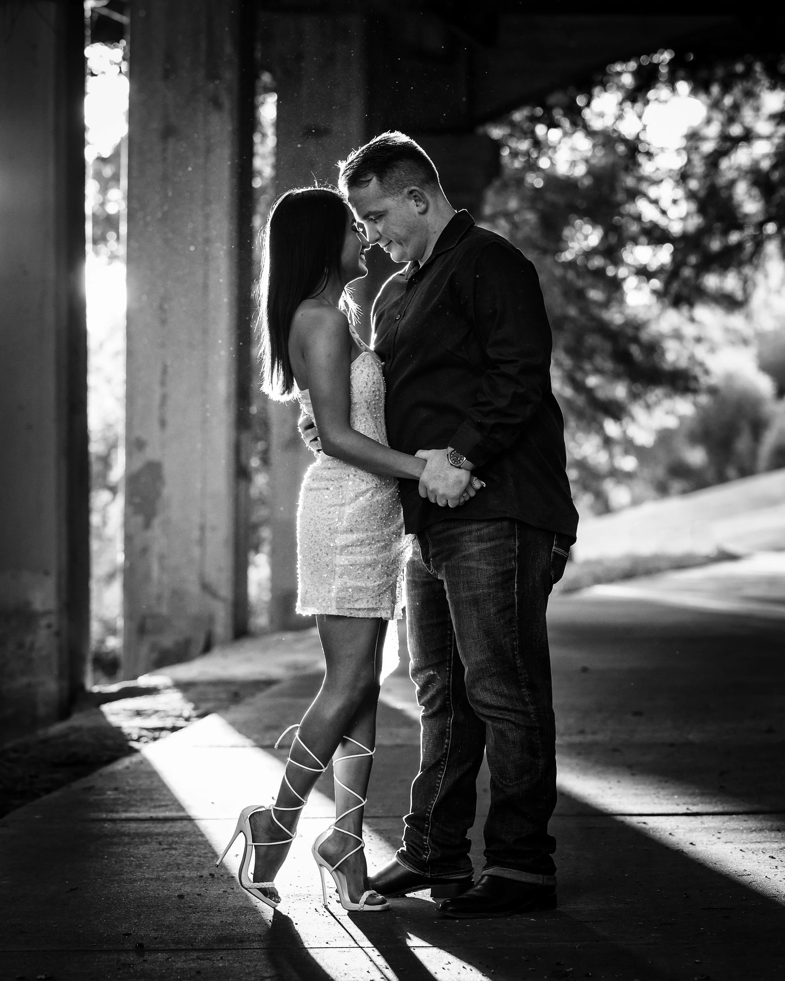 Black and white photo of a couple standing very close, noses almost touching, with sunlight creating a halo effect on their hair, outdoors near trees.