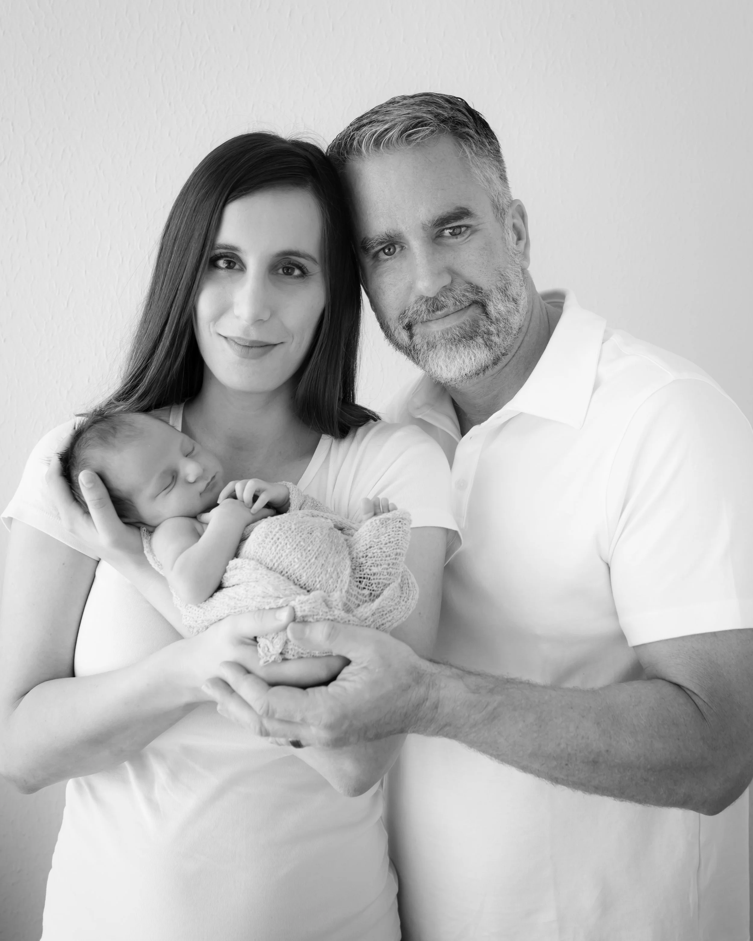 A black and white photo of a family of three with a newborn. The woman holds the baby, the man is close behind them, and they are all smiling. The scene appears to be indoors with a plain wall in the background.