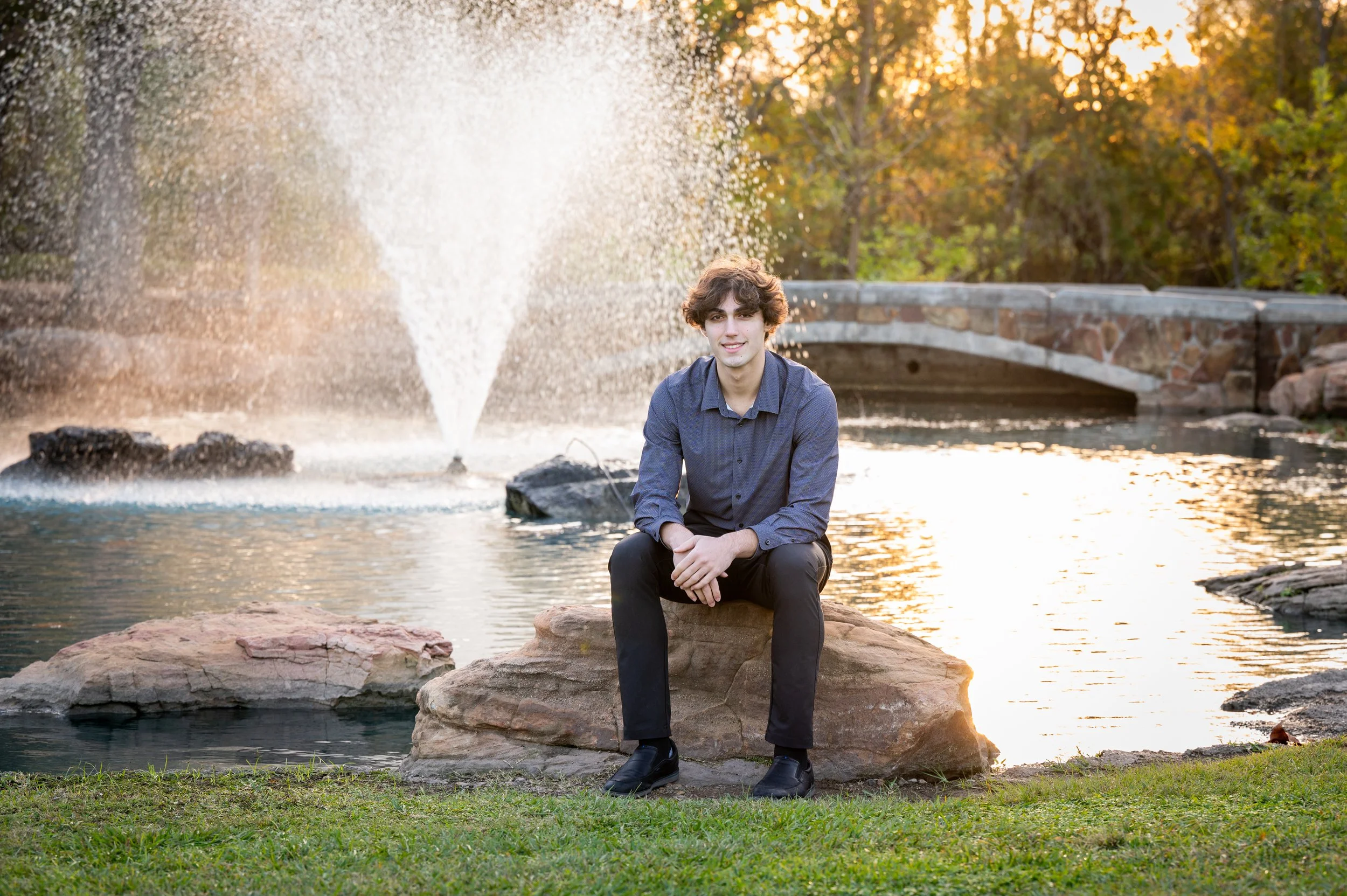 A young man with curly hair and a blue shirt sitting on a large rock near a body of water during sunset, with trees, a fountain and a stone bridge in the background.