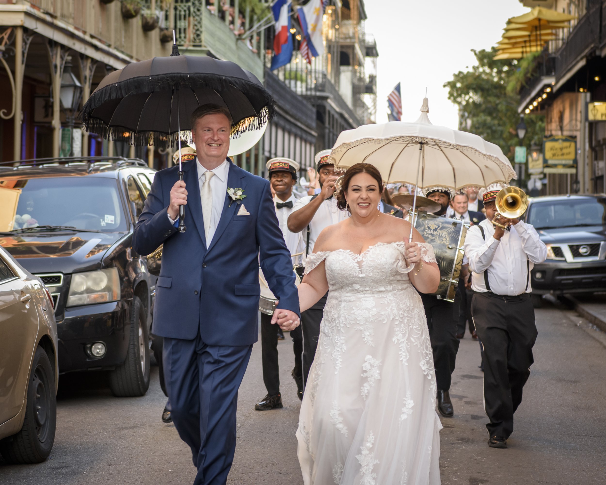 A bride and groom walking hand in hand during a second line wedding parade on a city street in New Orleans, surrounded by musicians and onlookers, with American flags and buildings in the background.