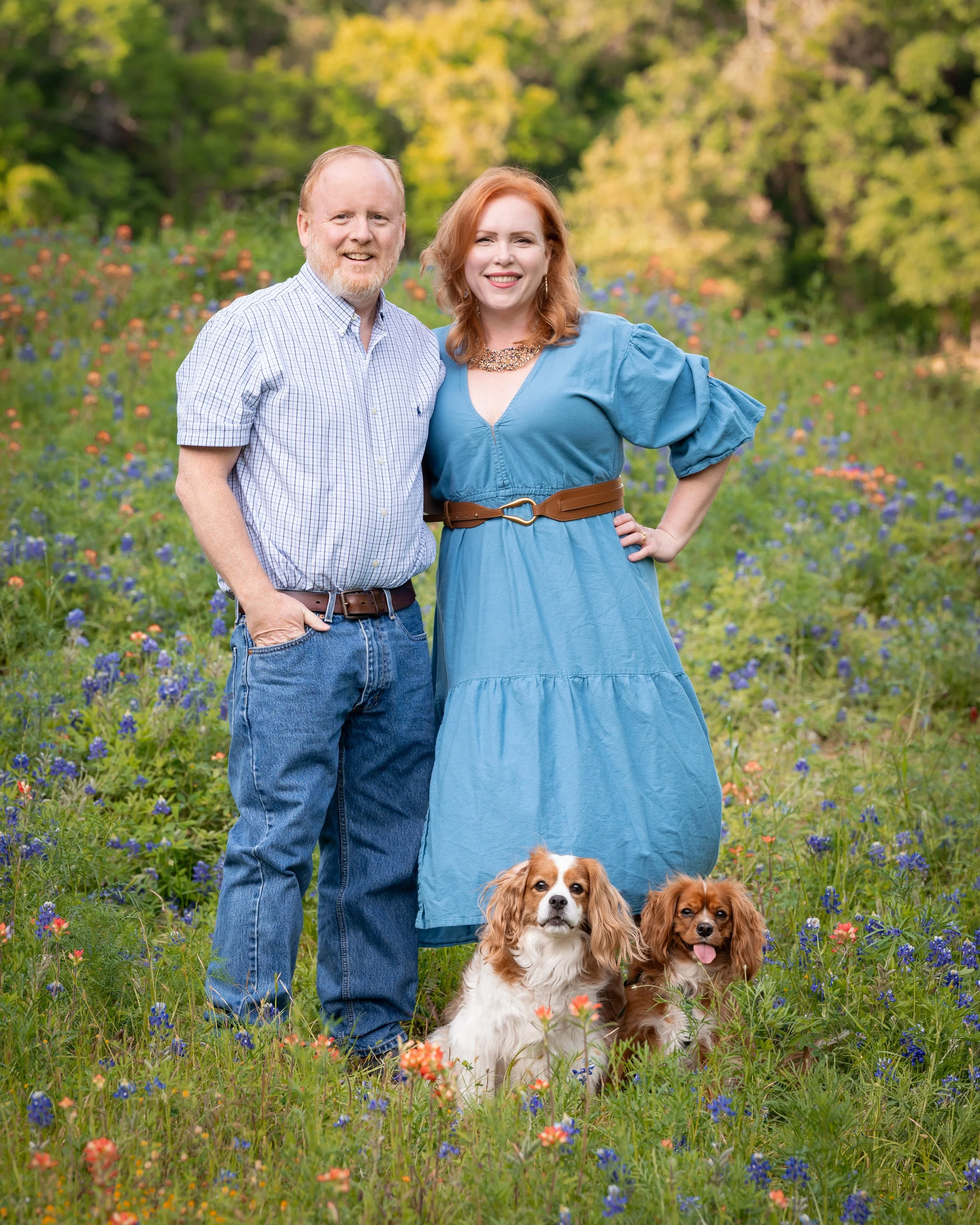 A man and woman stand in a field of colorful bluebonnets with two dogs, smiling at the camera on a sunny day with green trees in the background.