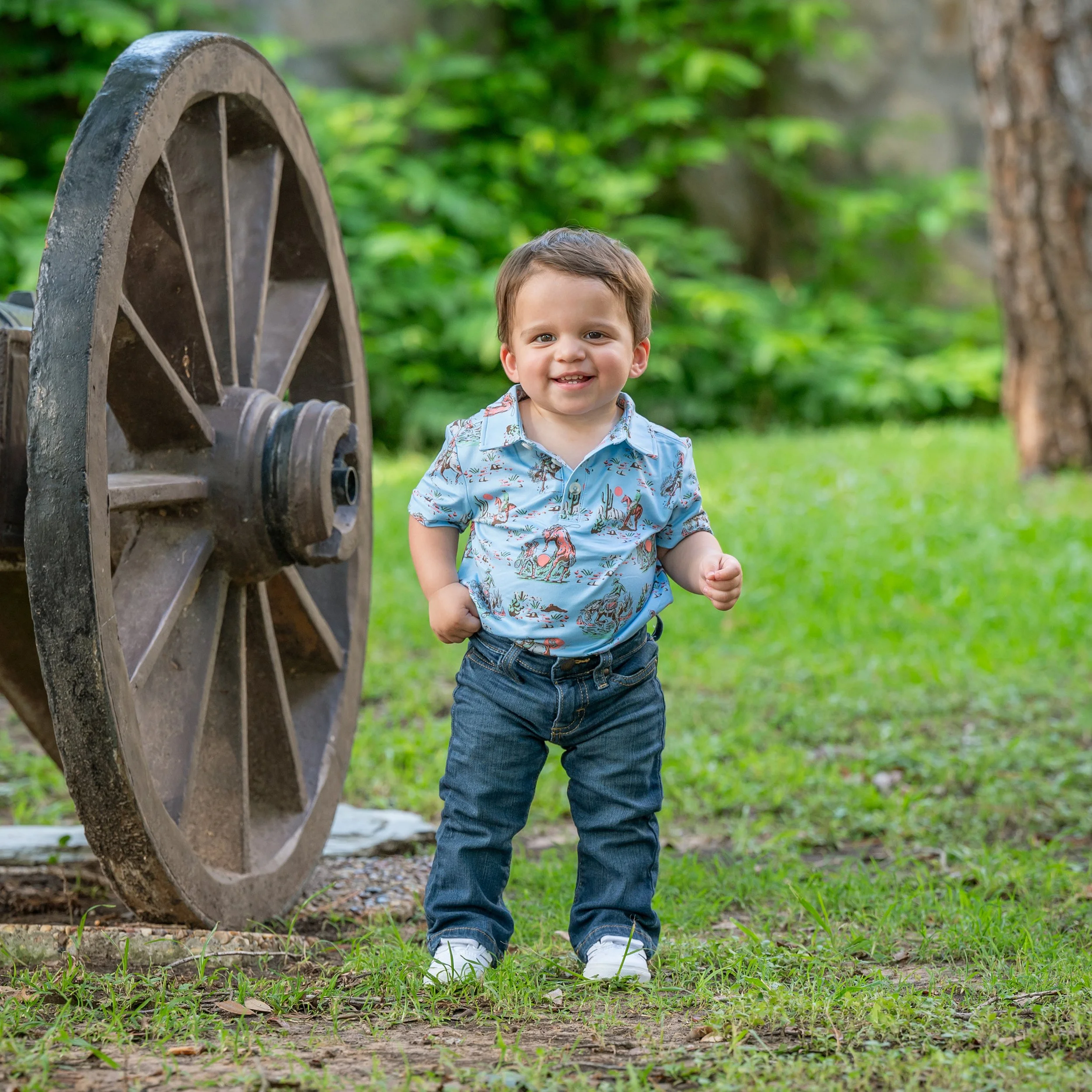 A young boy smiling and running outdoors next to a large wooden wagon wheel with a background of green trees and grass.