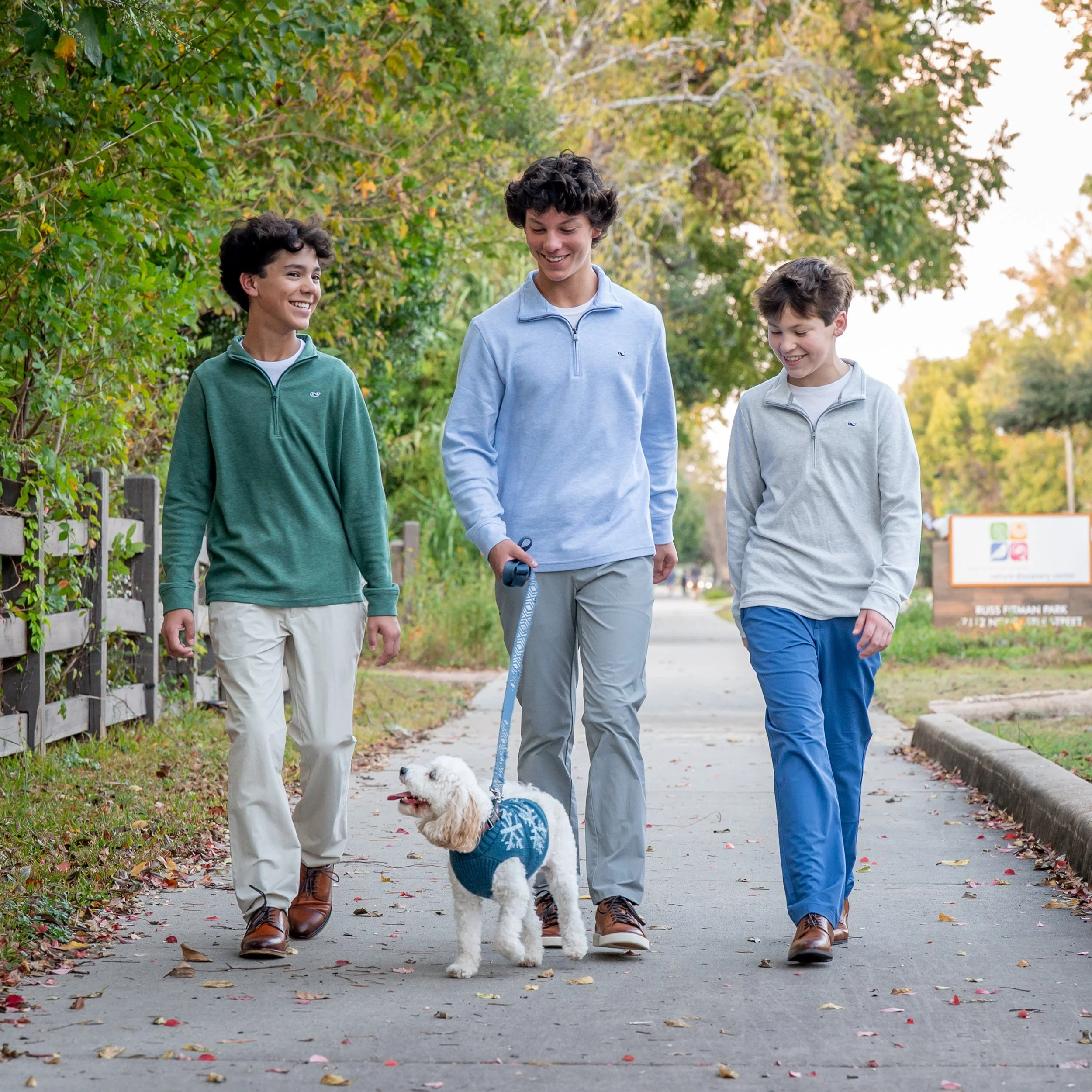 Three young men walking outdoors with a small white dog on a leash during fall, surrounded by trees with green and yellow leaves.