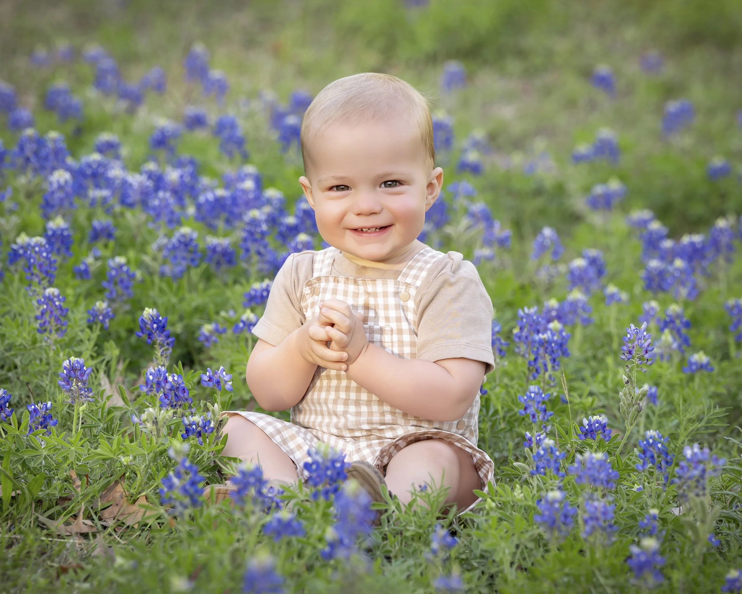 A young child with light hair, smiling, sitting among green grass and bluebonnet flowers, wearing a beige and white checkered outfit.