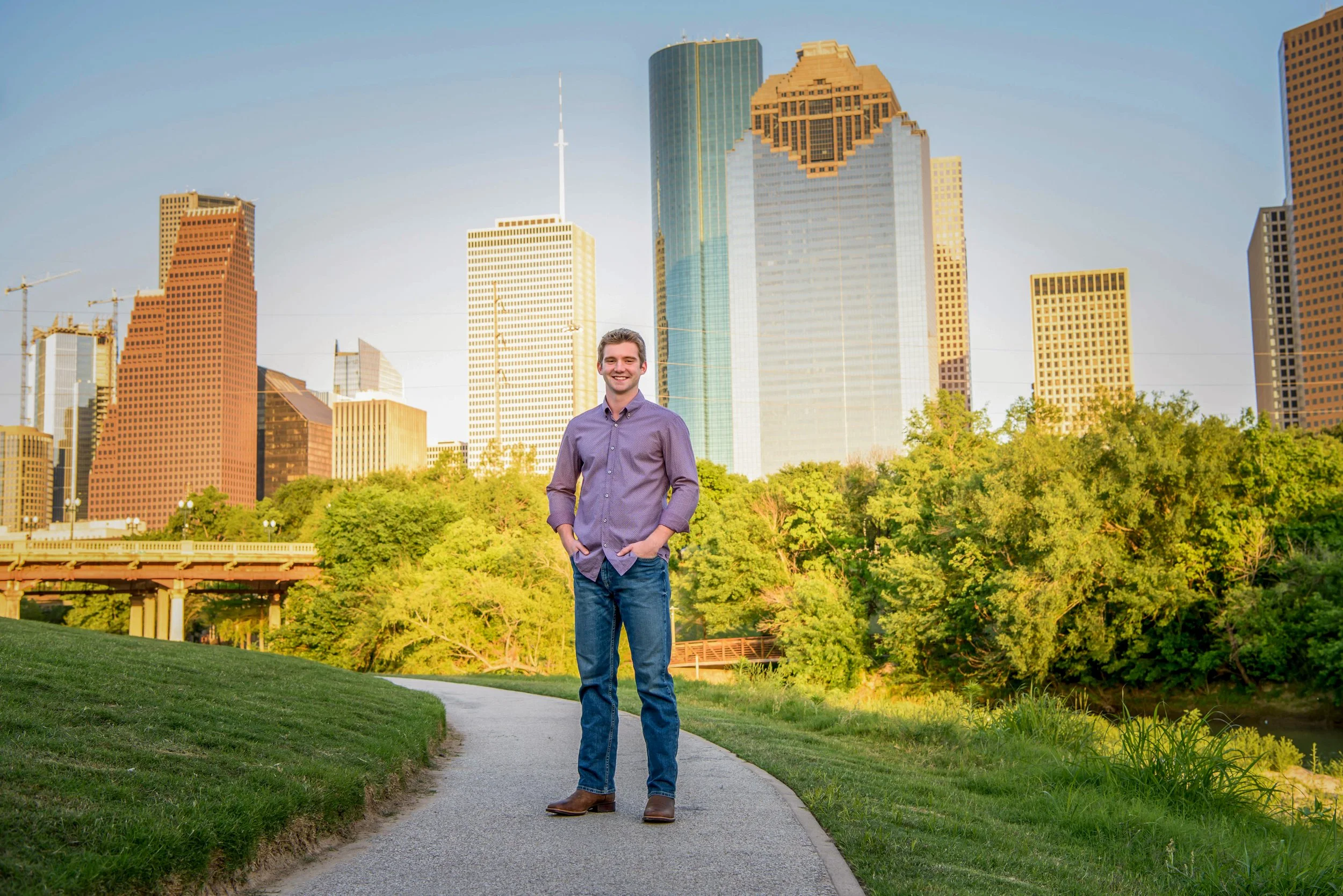 A senior guy in a purple shirt and jeans standing on a park trail with a city skyline of tall buildings in the background during late afternoon or early evening.