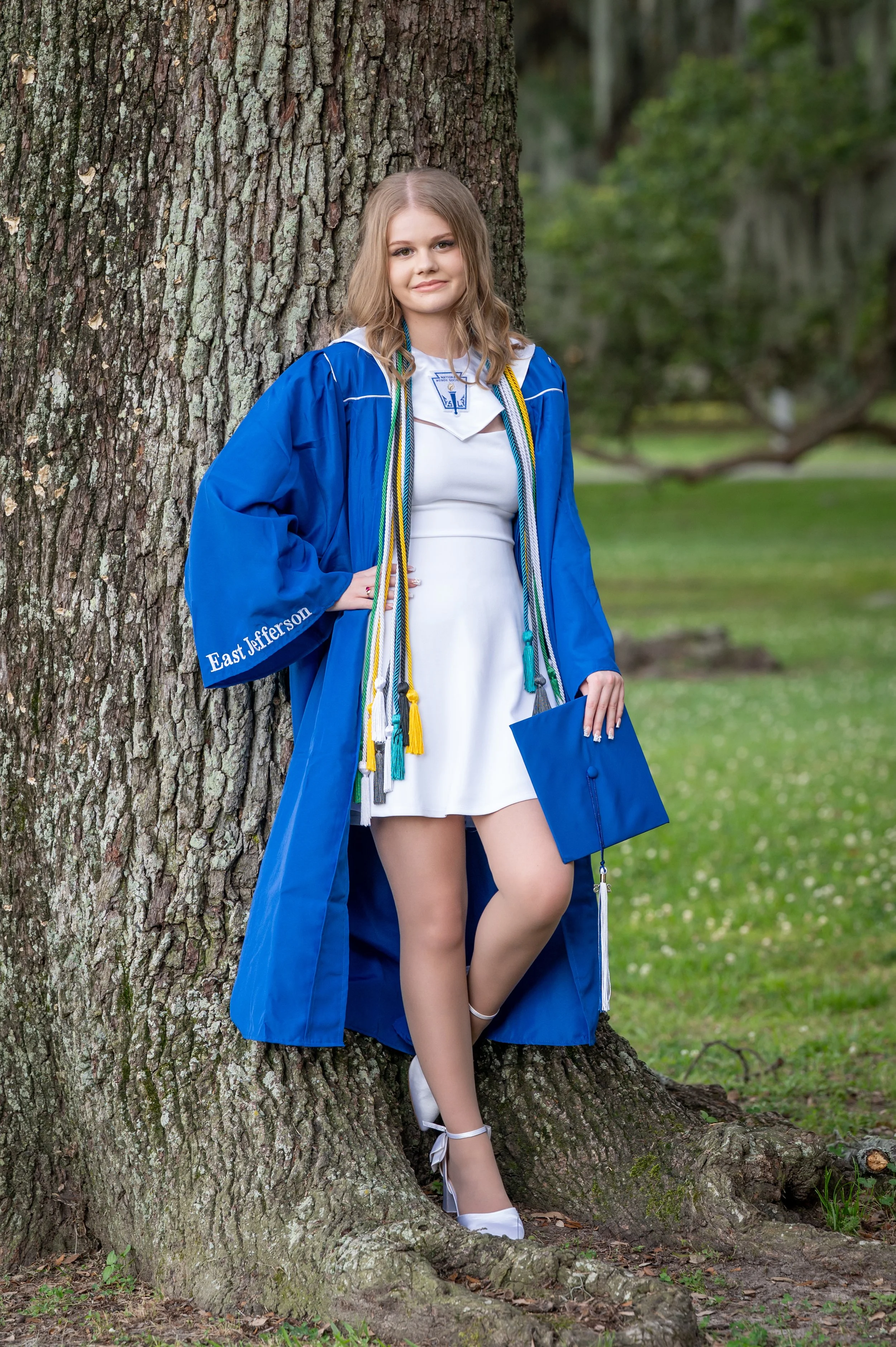 A young woman in a white dress and blue graduation gown with the words "East Jefferson" on the sleeve, standing against a large tree in a park-like setting, holding a blue diploma cover, with several cords around her neck, smiling at the camera.