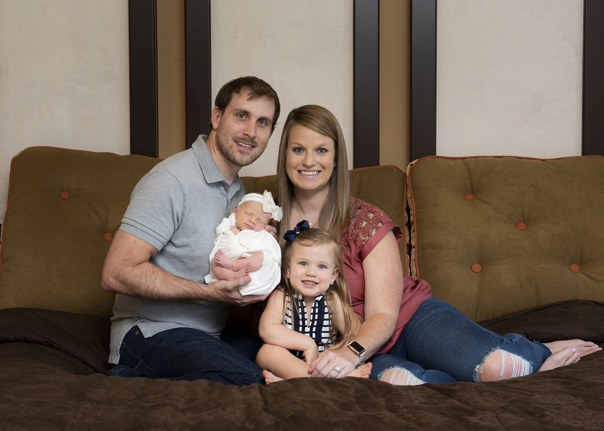 A happy family sitting on a sofa in a hotel room, holding a newborn baby and a young girl with long blonde hair, all smiling at the camera.