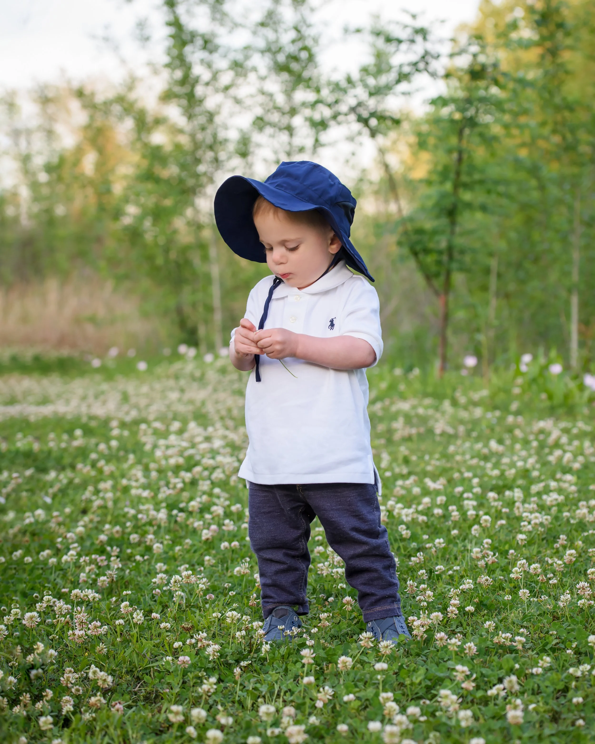 A young boy wearing a white polo shirt, navy hat, and dark jeans stands in a field of white and pink clover flowers, with green trees and a light sky in the background.