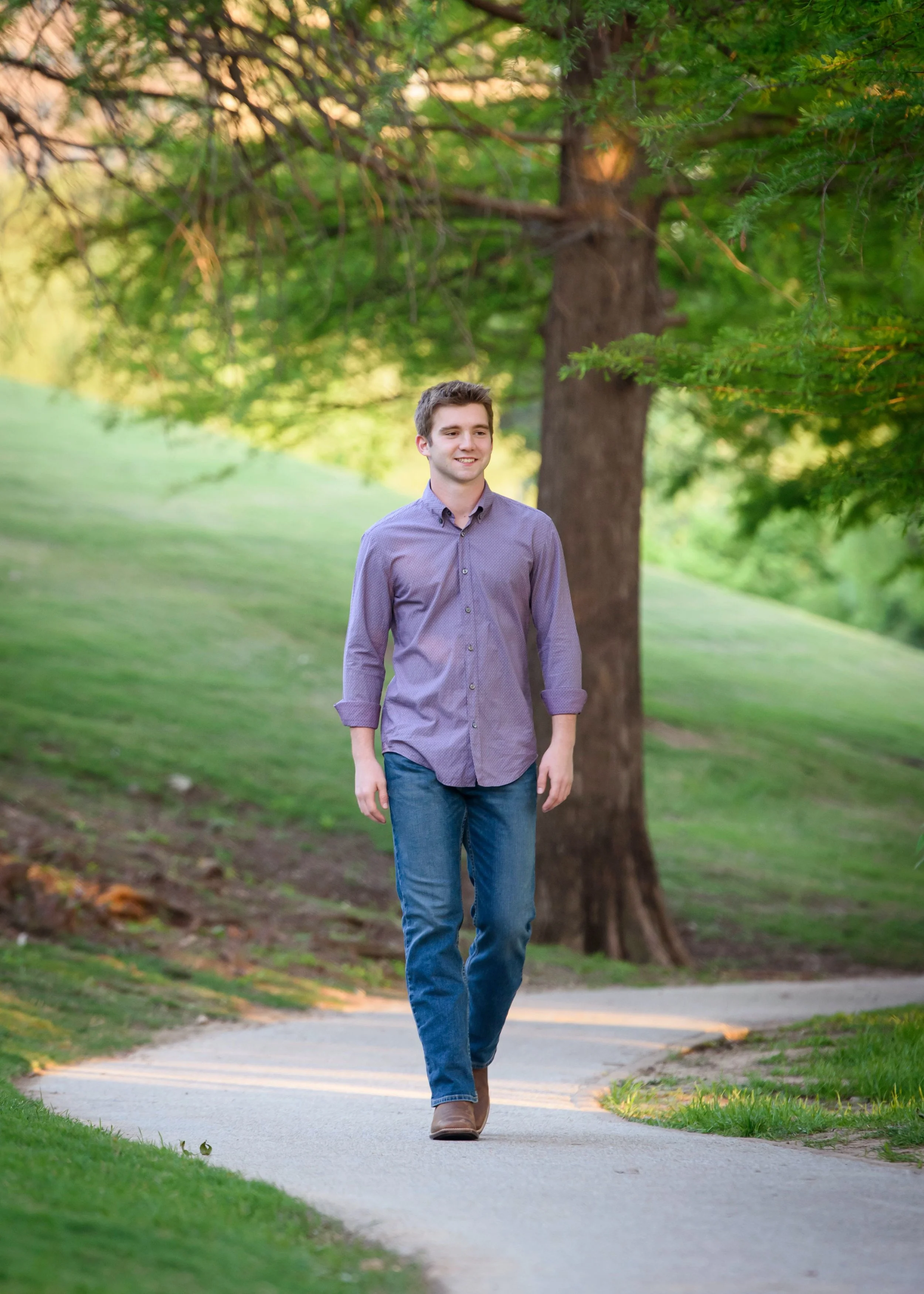 A senior guy walking on a paved path through a park with green grass and trees, smiling and looking to his left.