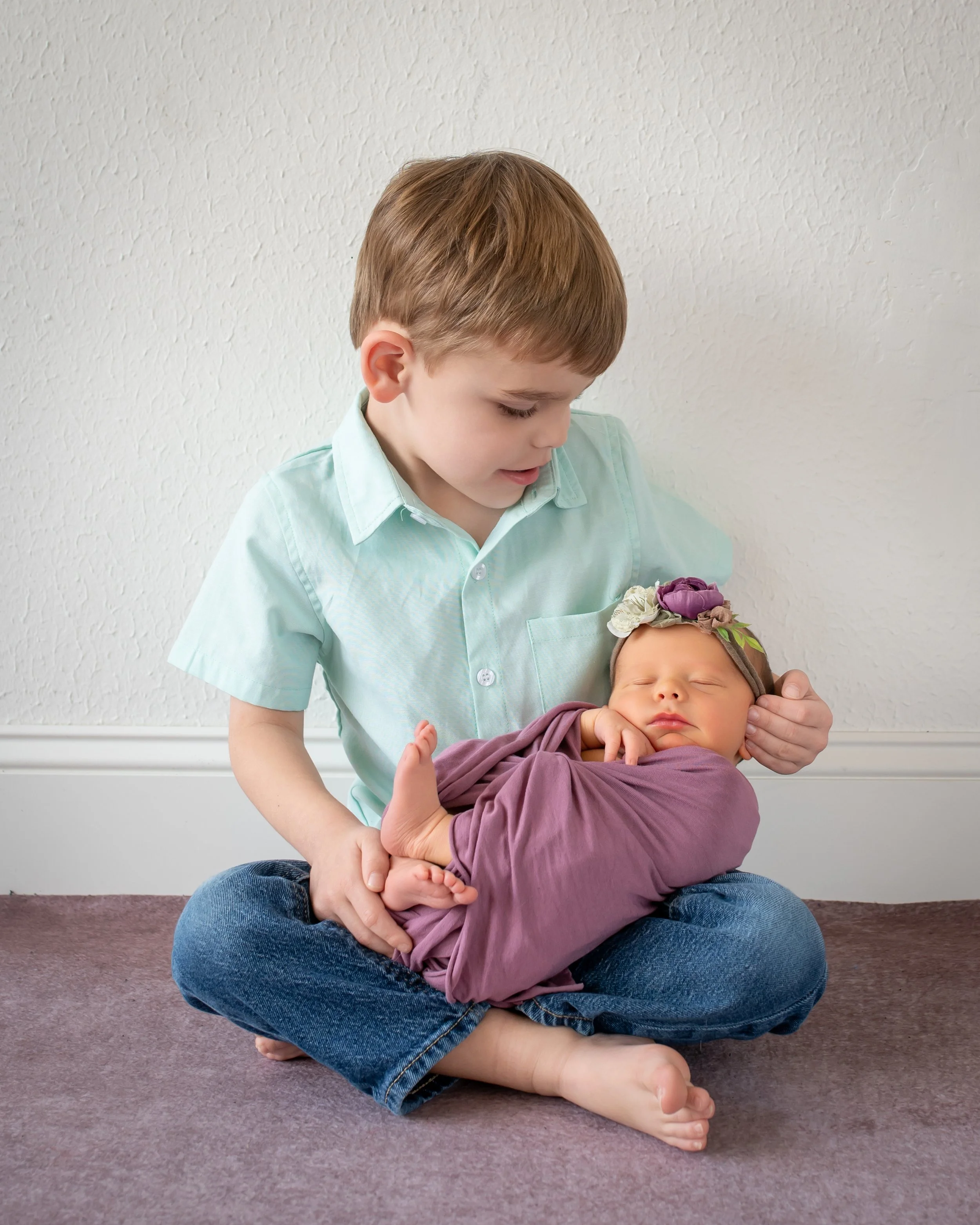 A young boy with brown hair and light skin sitting cross-legged on the carpeted floor, holding a sleeping newborn girl in his arms, both in front of a plain white wall.