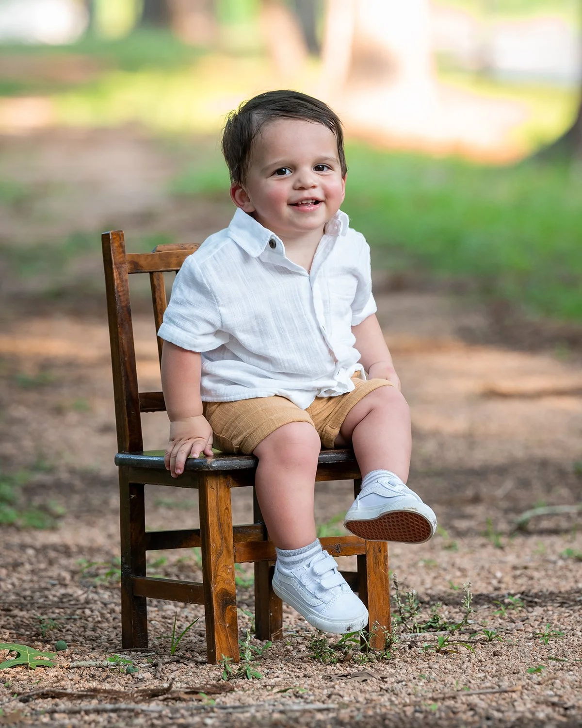 A young boy with dark hair, wearing a white shirt, khaki shorts, and white sneakers, sitting on a small wooden chair outdoors with a blurred green and brown background.