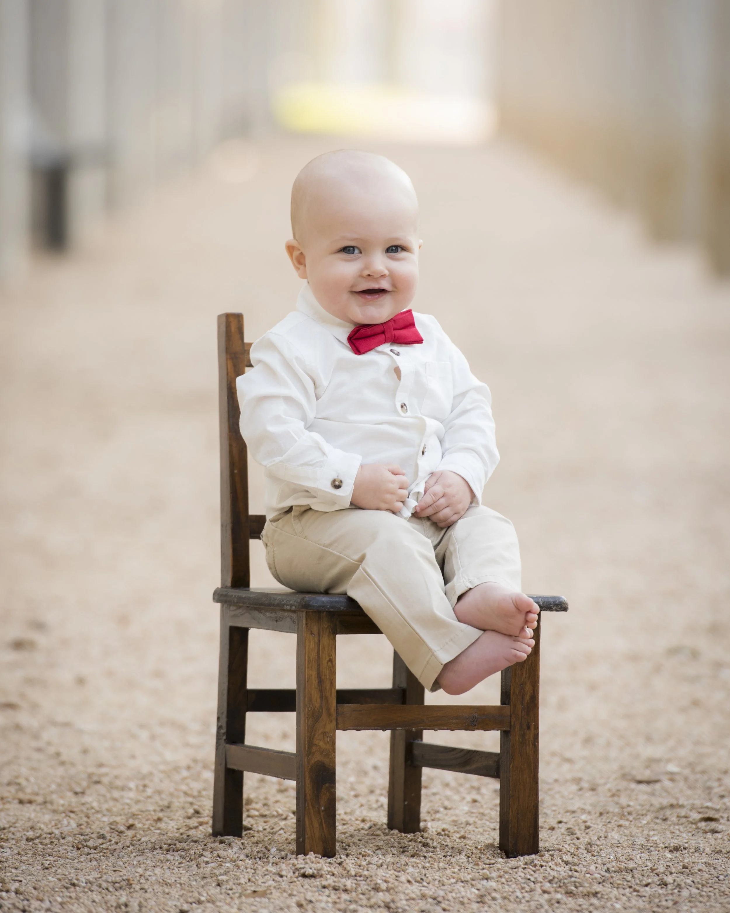 A smiling baby boy with a bald head, wearing a white shirt, red bow tie, and beige pants, sitting barefoot on an old wooden chair outdoors on gravel ground.
