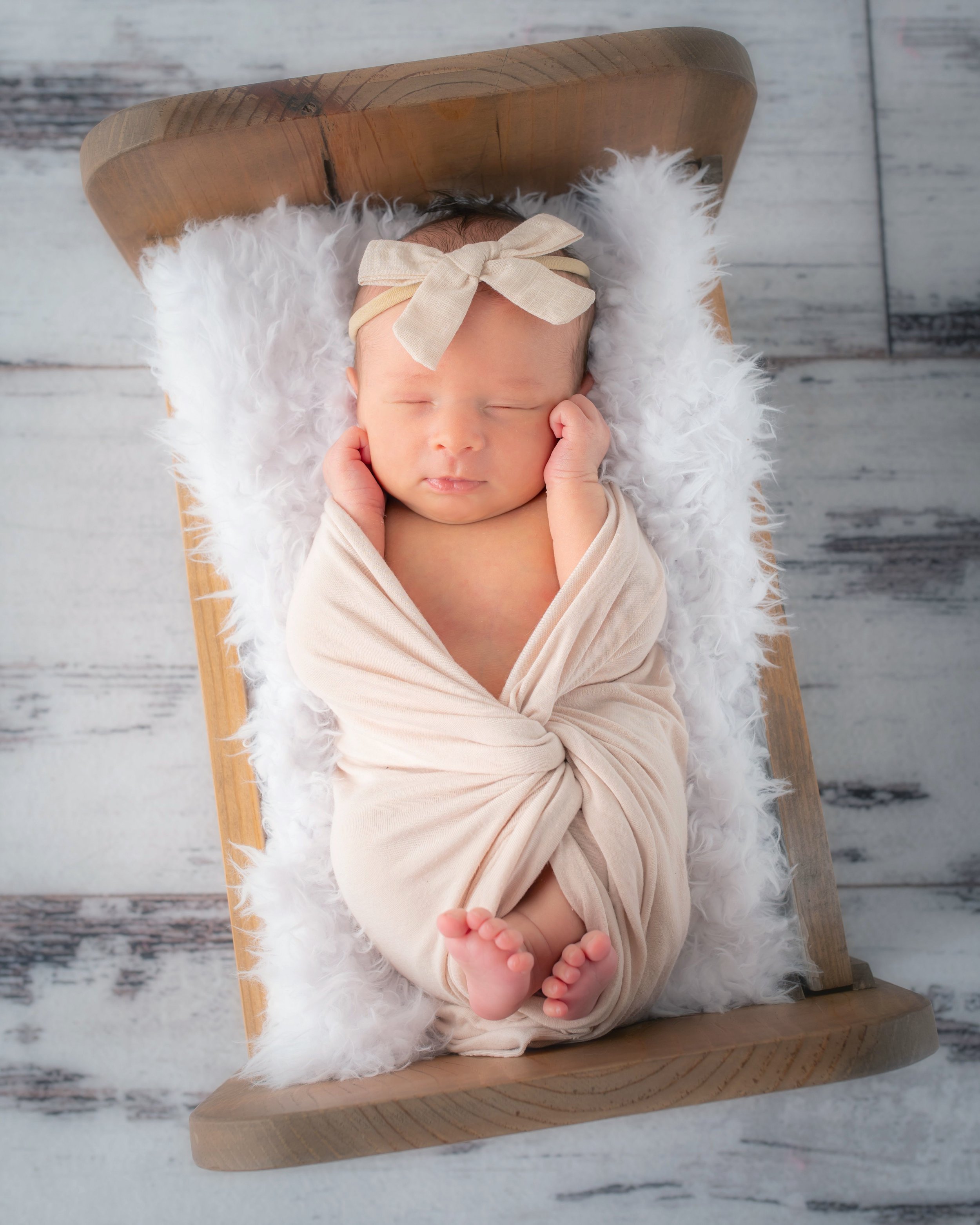 Newborn baby sleeping on a wooden bench wrapped in a beige blanket with a beige bow headband, surrounded by a white fluffy blanket, with a rustic wooden background.