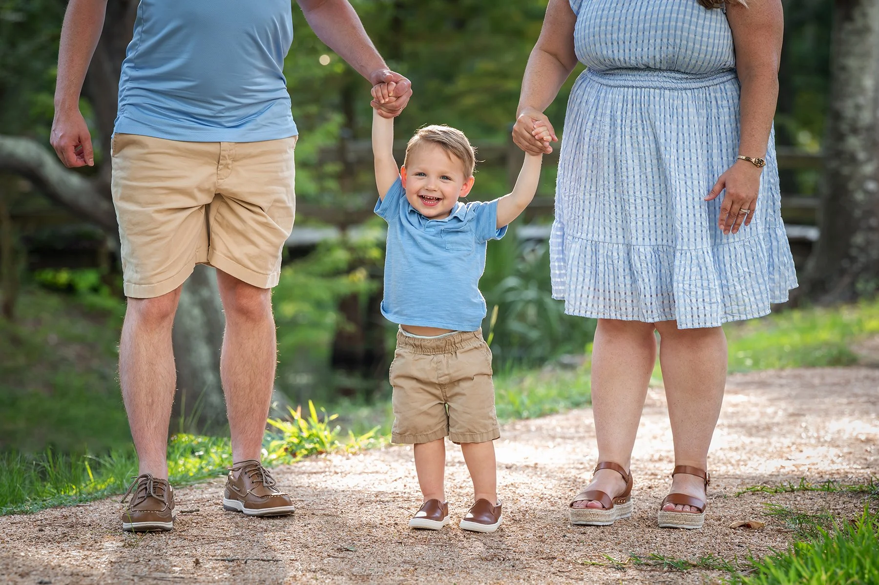 A young boy in a blue t-shirt and beige shorts holding hands with two adults, likely his parents, walking outdoors on a sunny day surrounded by greenery.