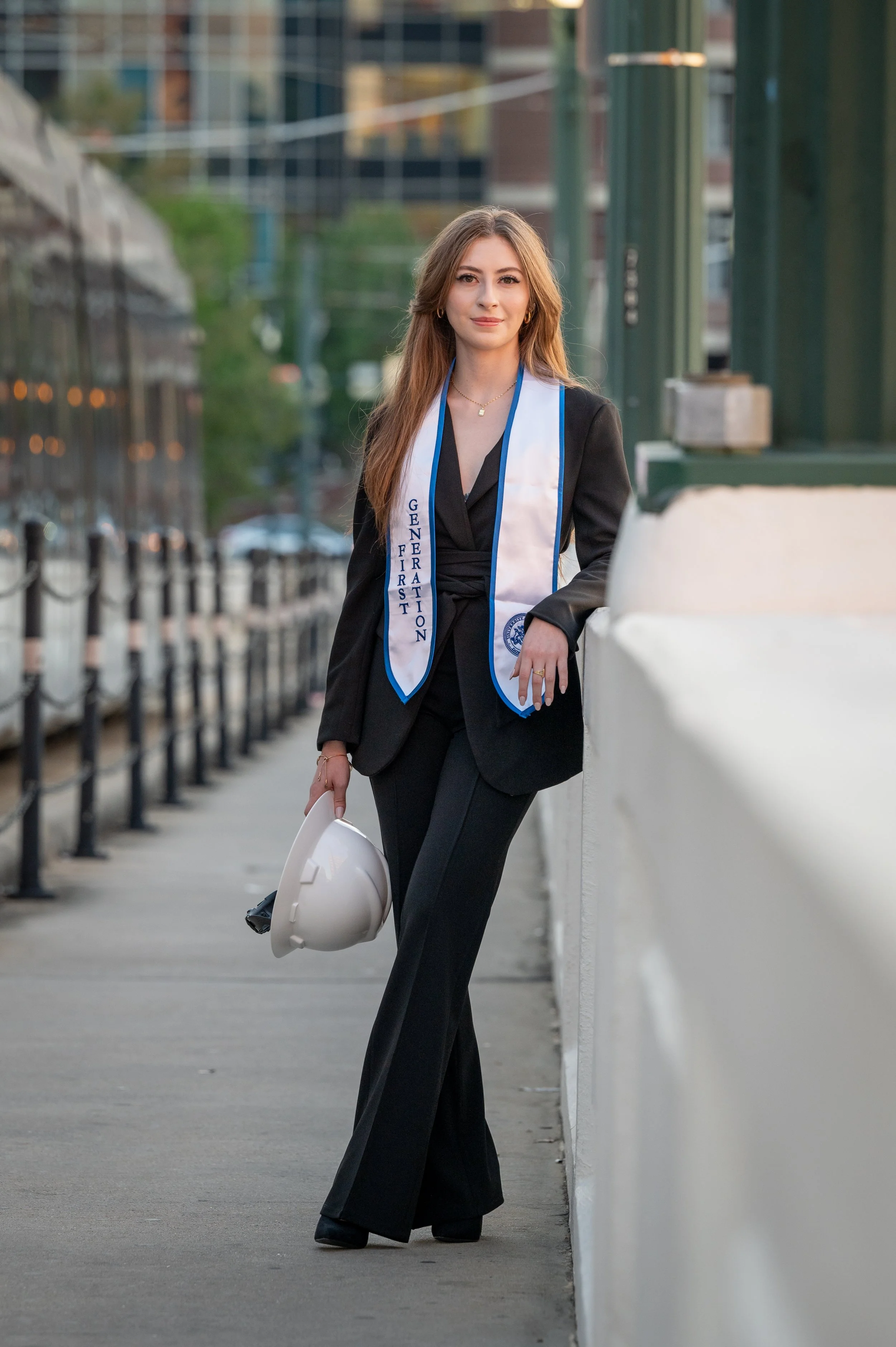 A college graduate with long brown hair stands outdoors on a city sidewalk, wearing a black blazer and pants, holding a white hard hat, and smiling slightly. She has a graduation sash that reads 'GENDER FIRST' and the university's emblem.