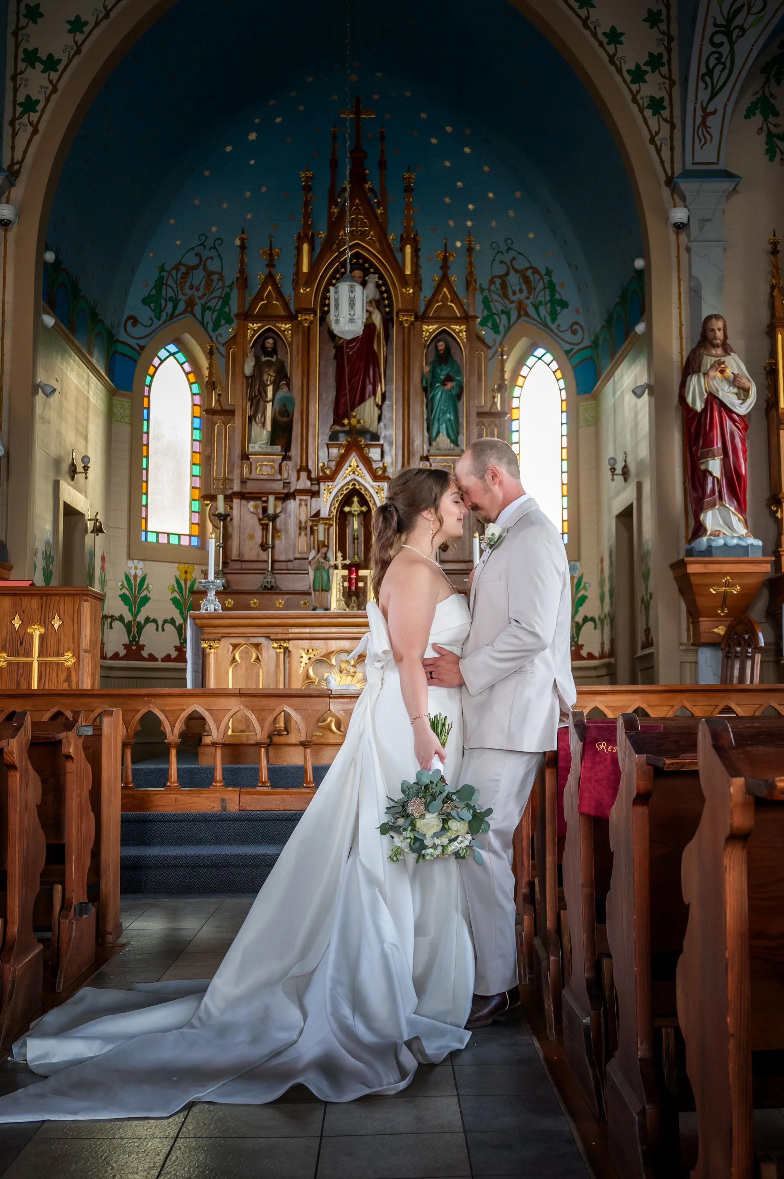A bride and groom standing close together in a painted church during their wedding ceremony, with church altars and religious statues in the background.